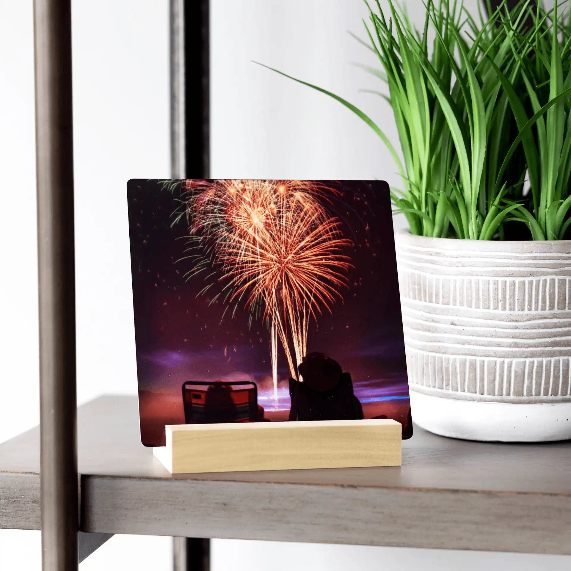 Metal desk print featuring colorful fireworks in a night sky above silhouetted chairs, displayed on a shelf beside a textured plant pot.