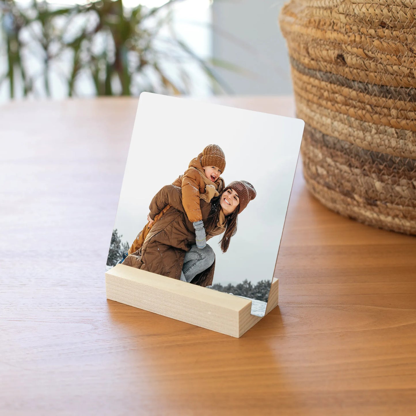 Metal desk print of a woman giving a child a piggyback ride in snow, displayed in a light wood block stand on a table.