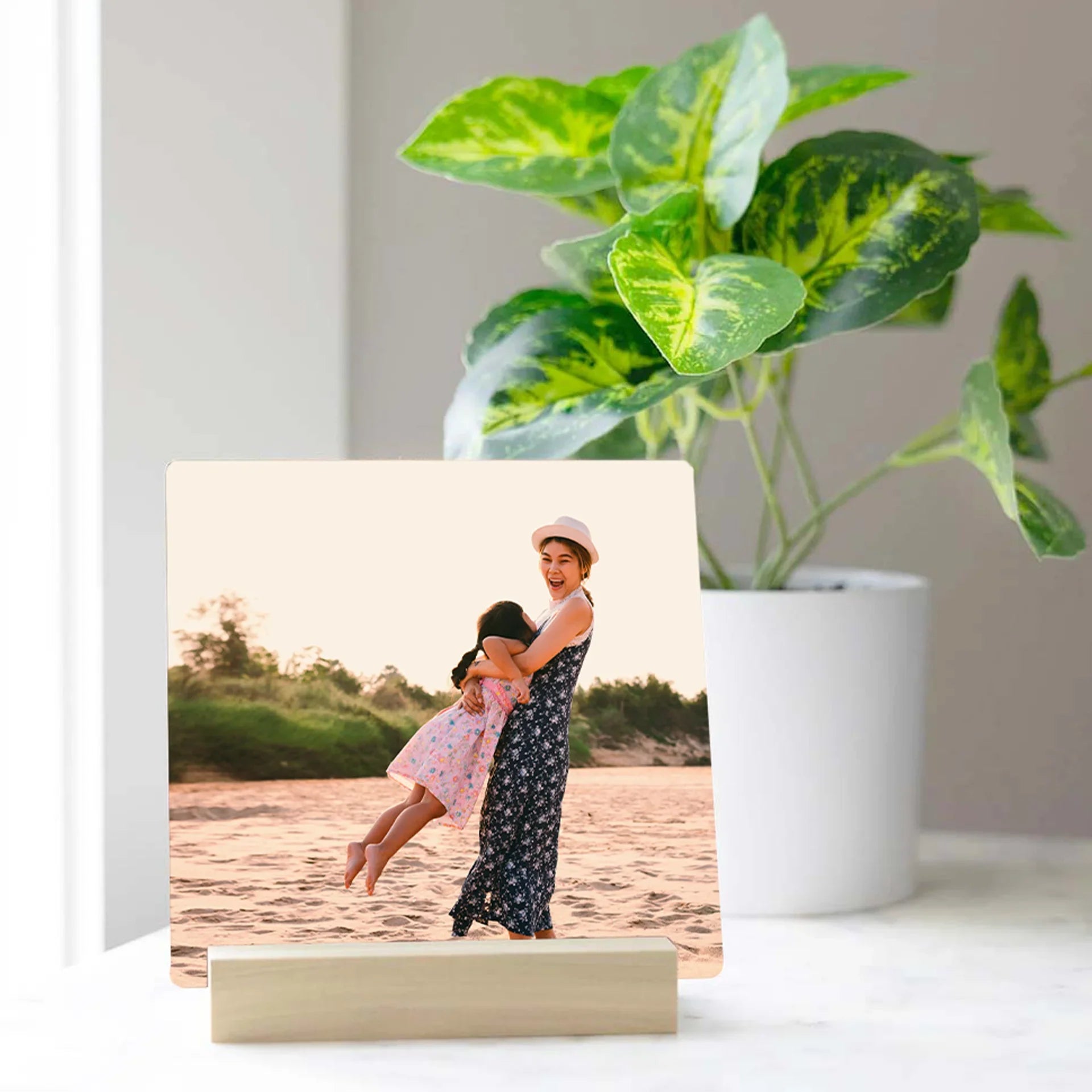 Metal desk print featuring a mother lifting her daughter at the beach, displayed on a white tabletop beside a green potted plant.
