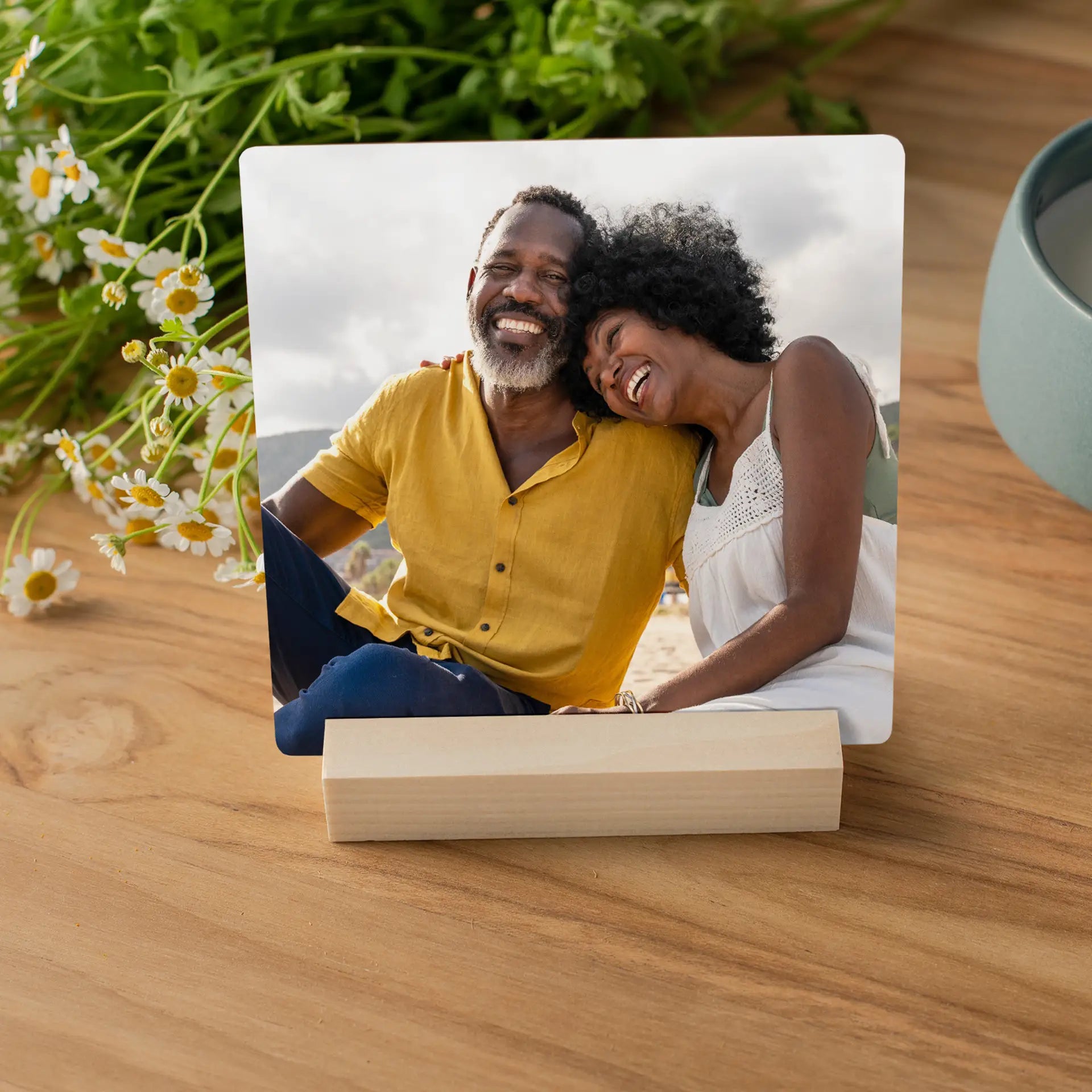 Metal desk print showing a smiling couple leaning together outdoors, displayed on a wooden surface with fresh flowers and a bowl nearby.