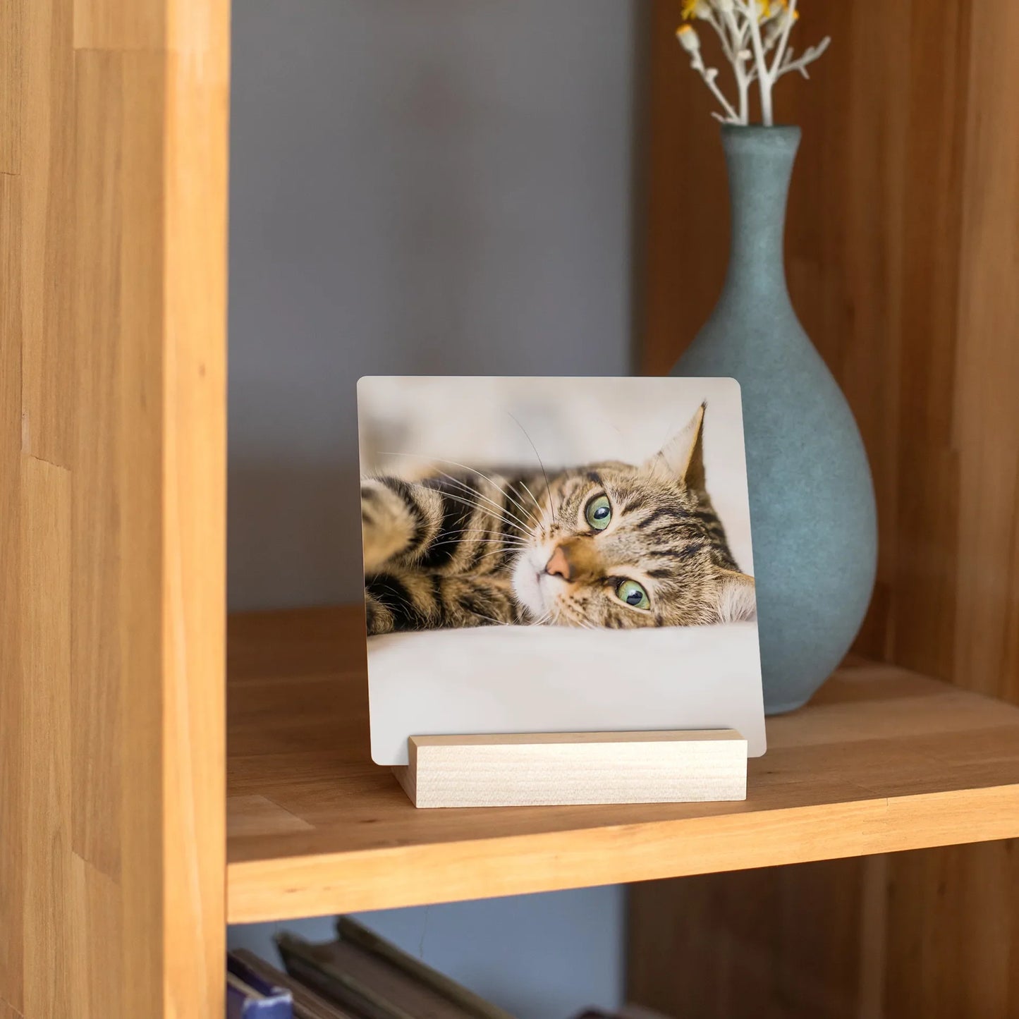 Metal desk print featuring a close-up of a tabby cat with green eyes, displayed on a wooden block stand on a shelf beside a ceramic vase.