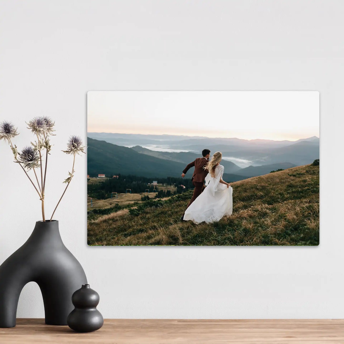Metal wall decor print featuring a couple in wedding attire walking across a grassy mountain hillside at sunset with layered landscape views.