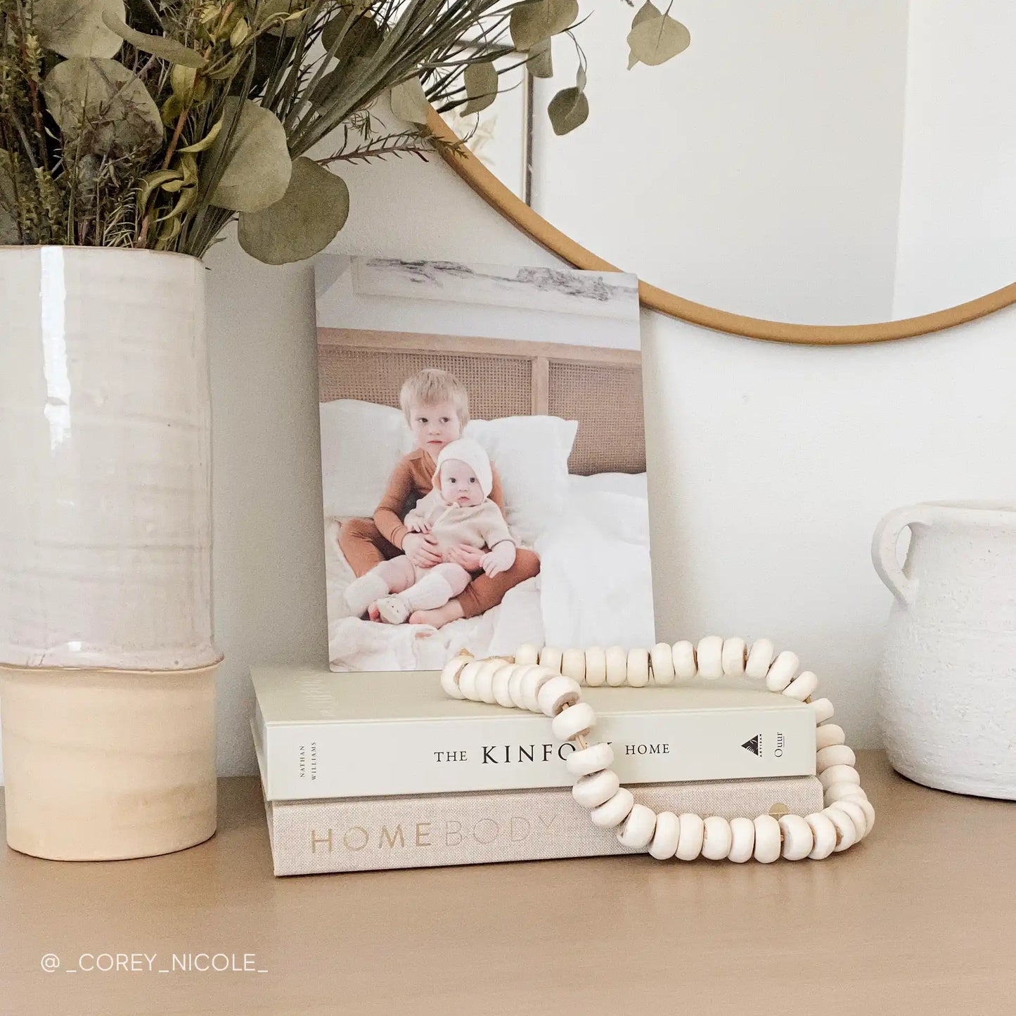 Metal wall print featuring two young children sitting together on a bed in a softly lit bedroom, displayed on stacked books beneath a round mirror.