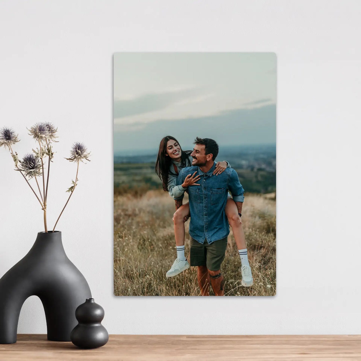 Metal wall decor print showing a couple in casual clothing with one person giving a piggyback ride in an open grassy field.