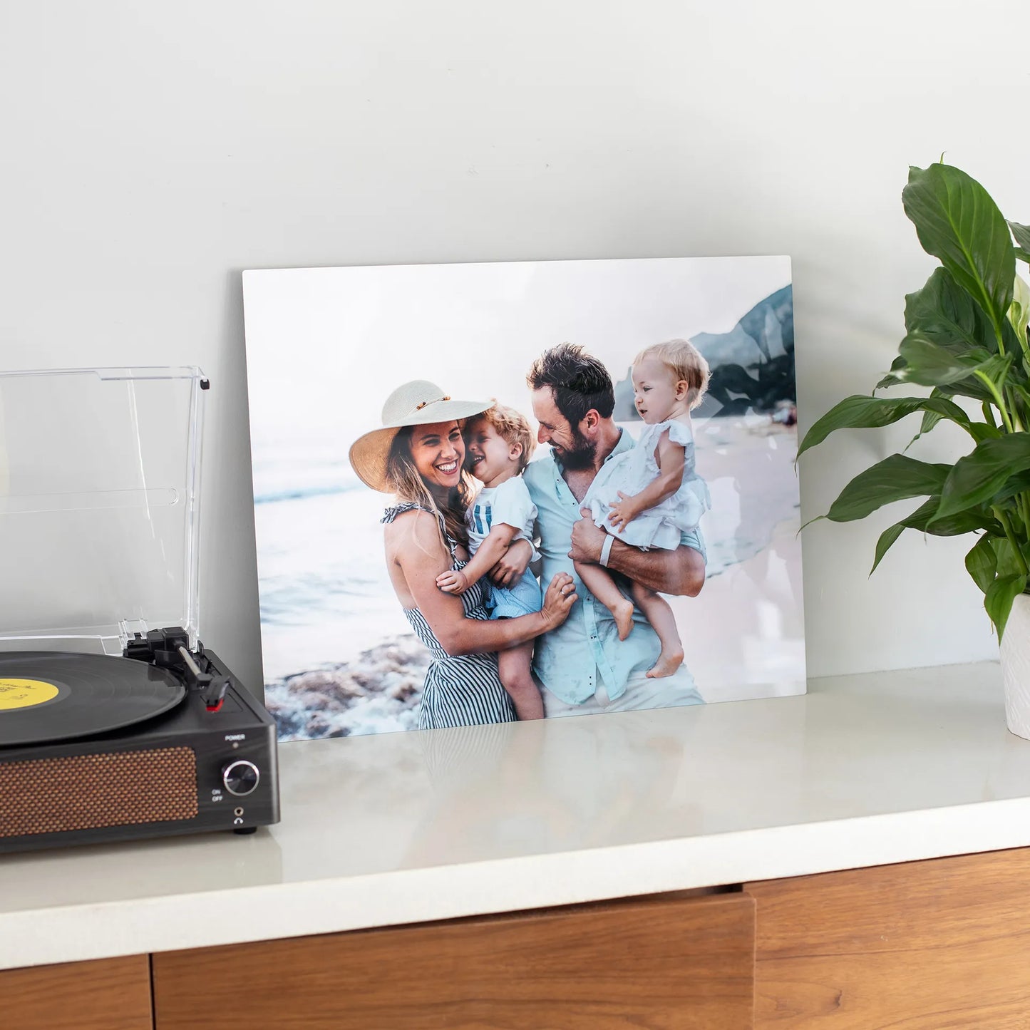 Metal wall print of a smiling family of four holding two young children on a beach, displayed on a console table beside a record player and plant.