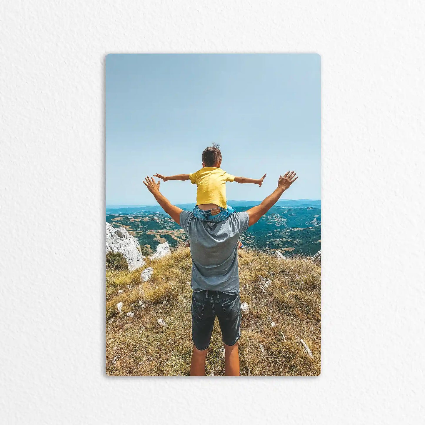 Metal wall decor print showing an adult carrying a child on their shoulders overlooking a wide mountain landscape under a clear sky.