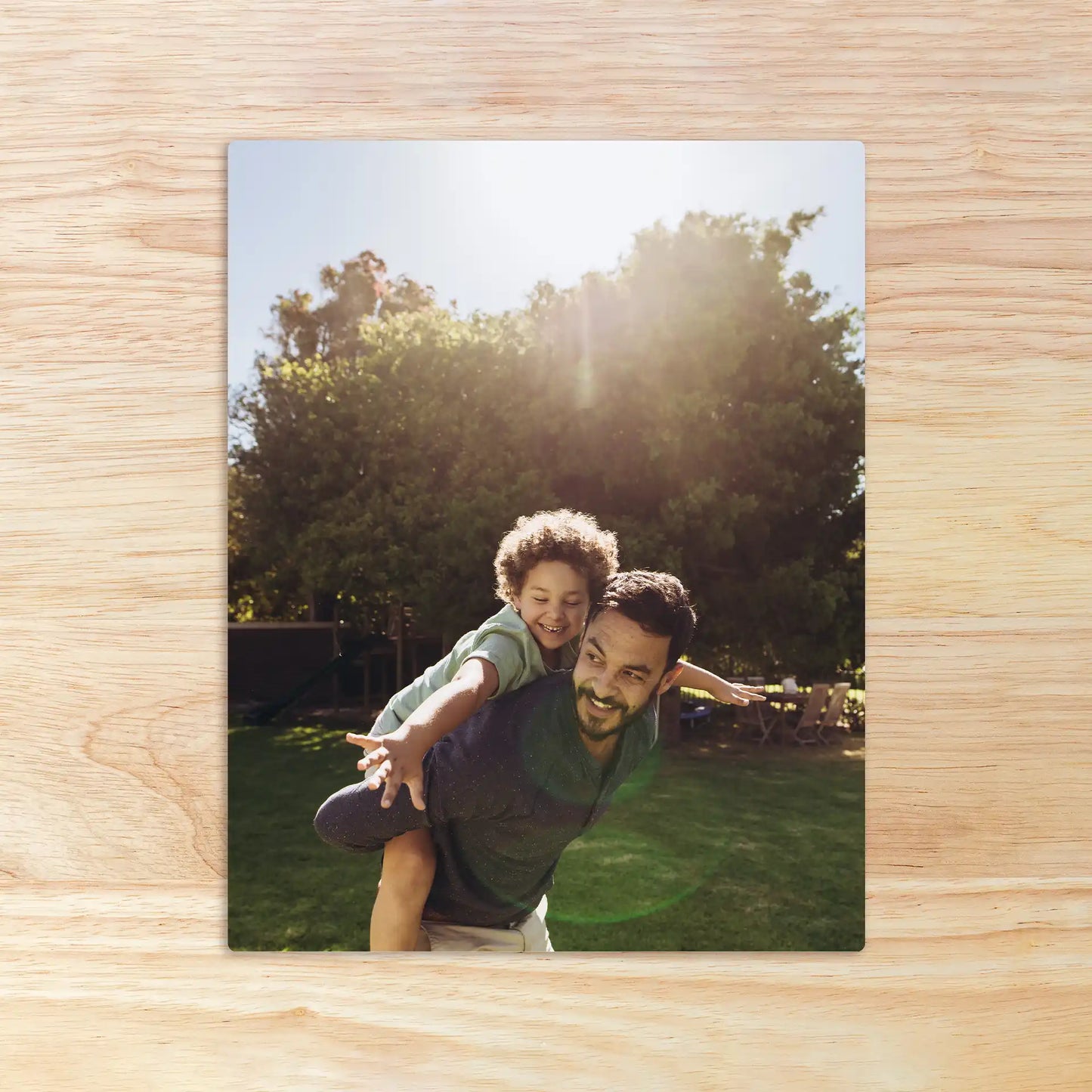 Metal wall decor print of an adult giving a child a piggyback ride in a sunlit backyard with trees and grass.