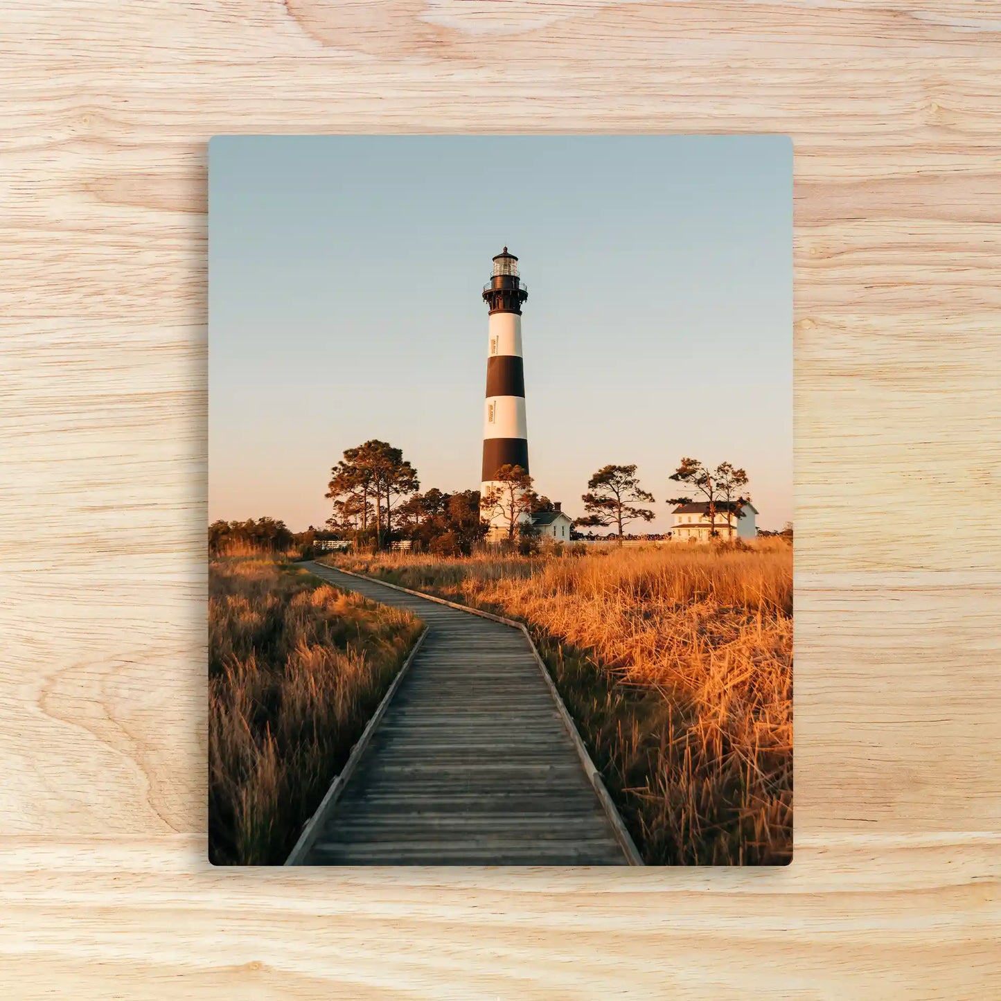 Metal wall decor print featuring a lighthouse with black and white stripes along a wooden boardwalk surrounded by tall grass.