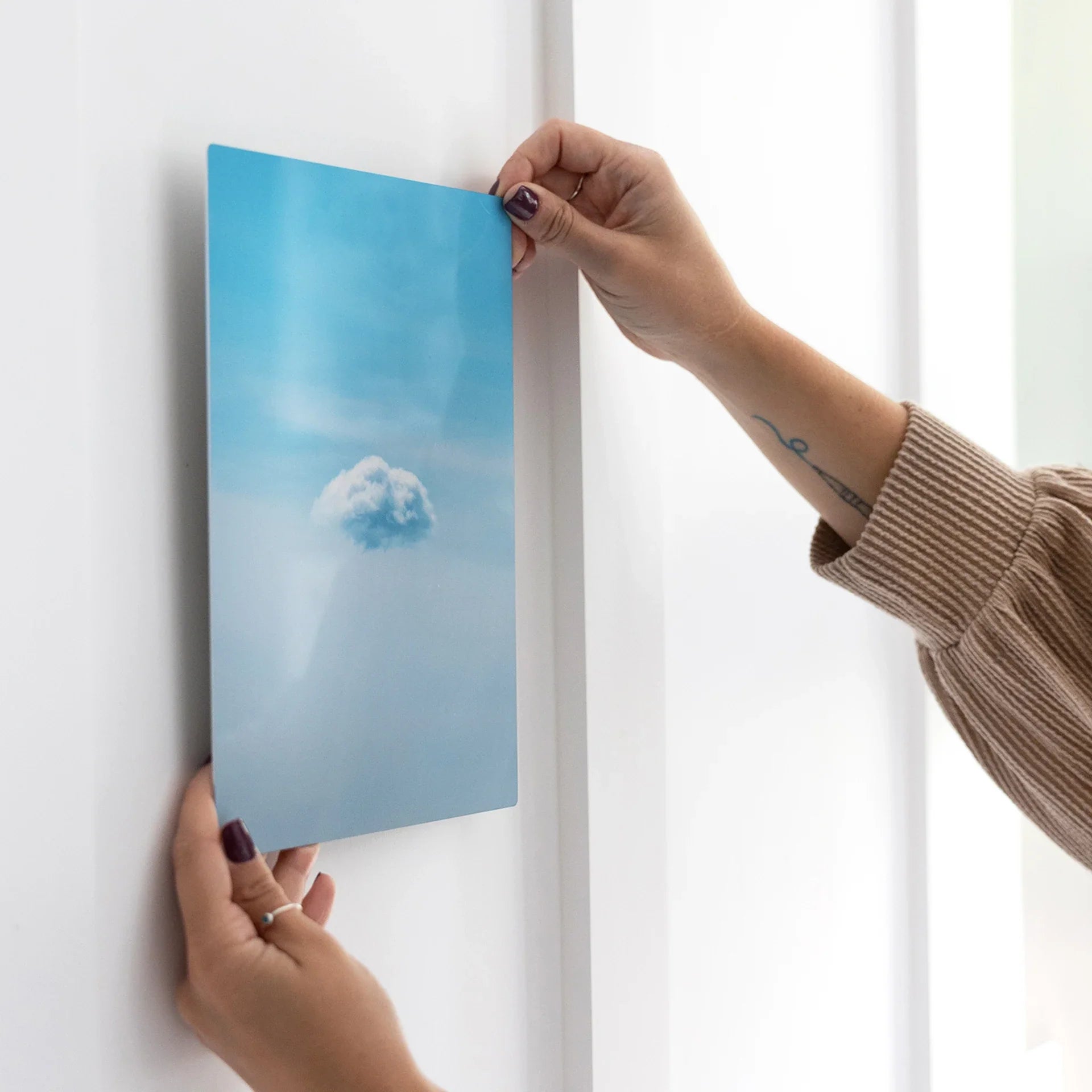 Metal wall print showing a single white cloud against a soft blue sky, being positioned on a white paneled wall.