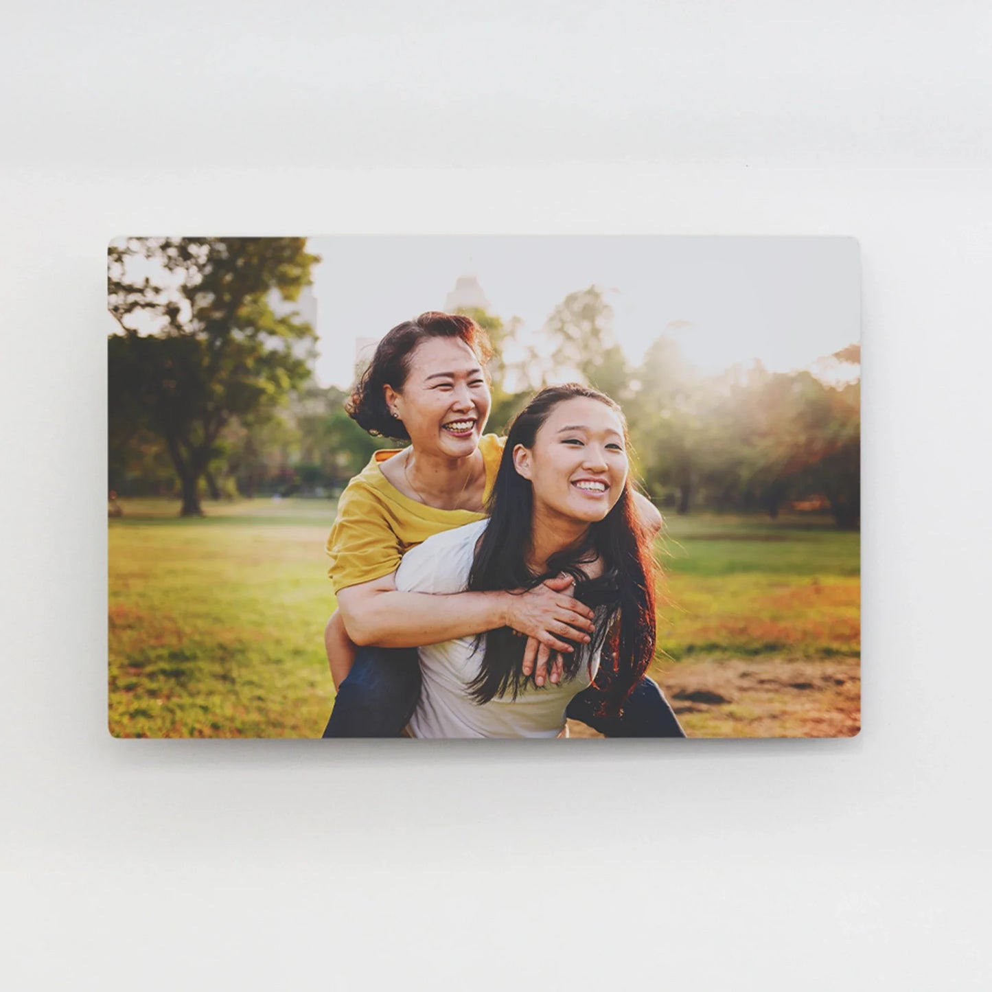 Metal wall decor print of two people smiling during a piggyback moment in a sunlit park with trees and grass in the background.