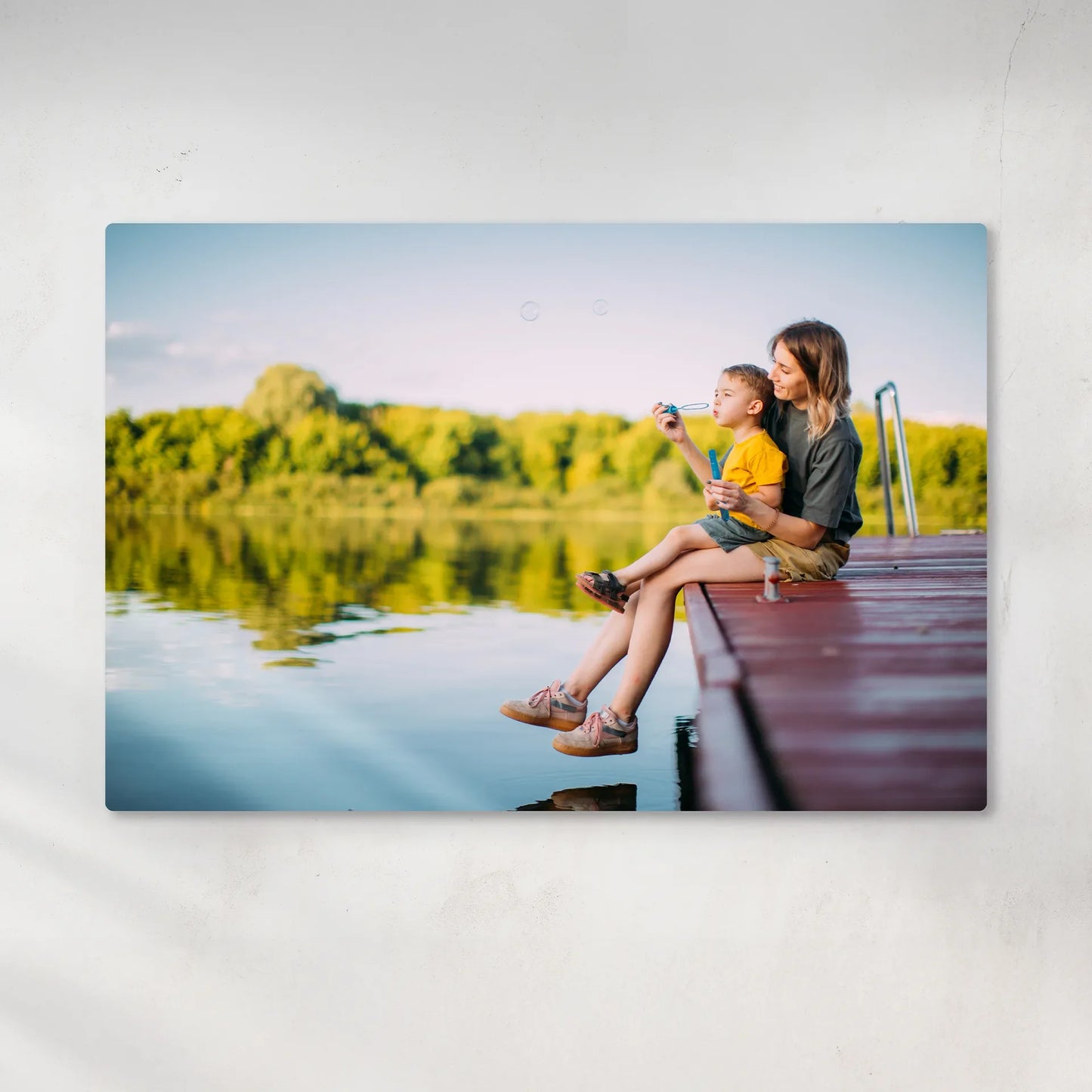 Metal wall decor print showing a parent and child sitting on a wooden dock by a calm lake with green trees reflected in the water.