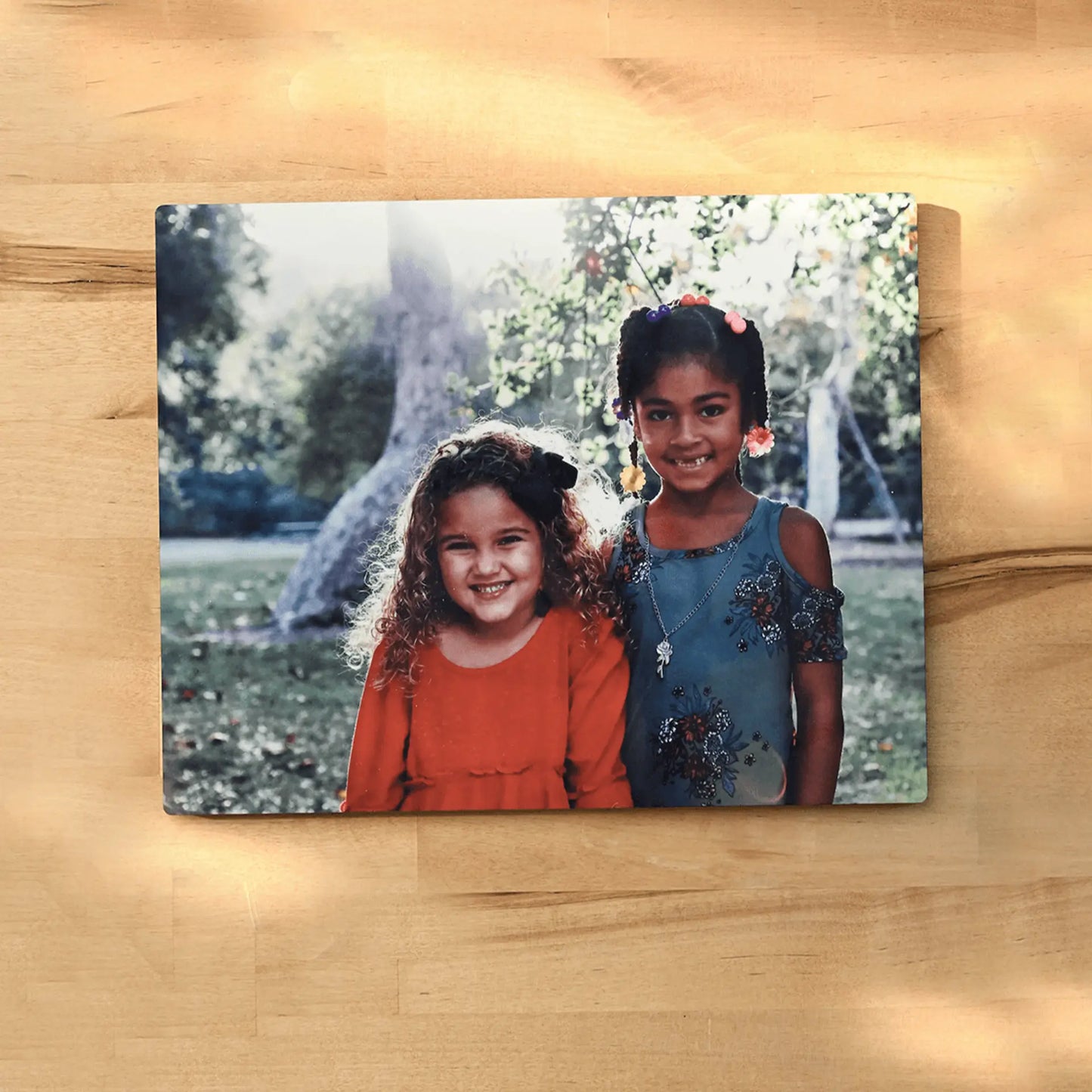 Metal wall print of two young girls smiling outdoors in a park setting, displayed flat against a light wood surface.
