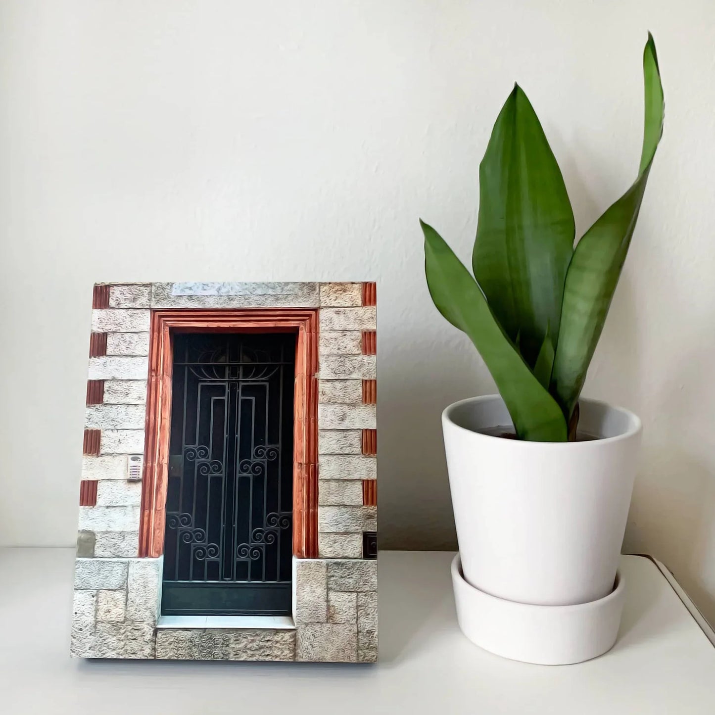 Mounted photo print of a stone building doorway with a decorative metal gate, displayed on a white surface beside a potted plant.