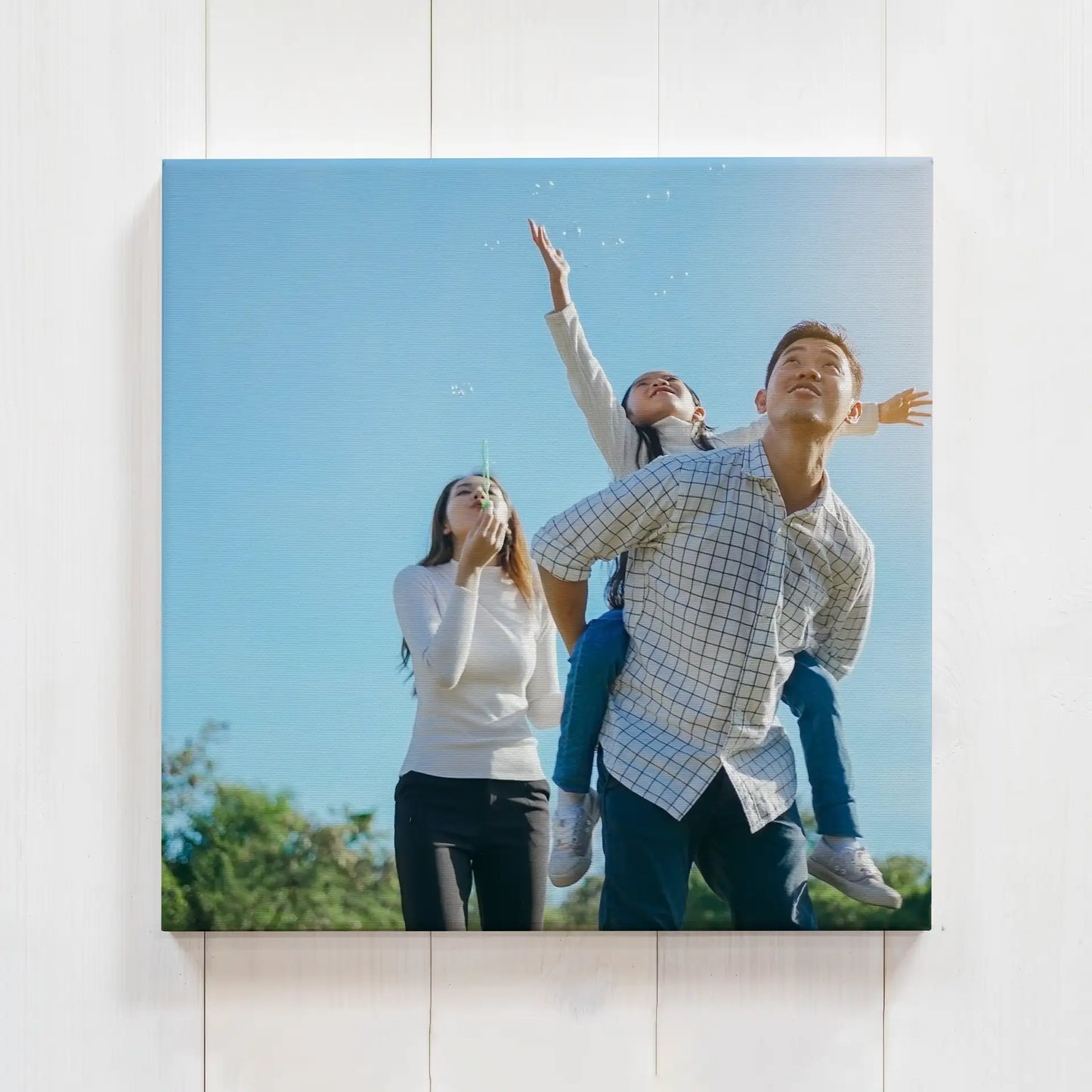 Mounted print with family outdoors under blue sky, child on shoulders reaching upward, bright natural lighting, frameless wall display.