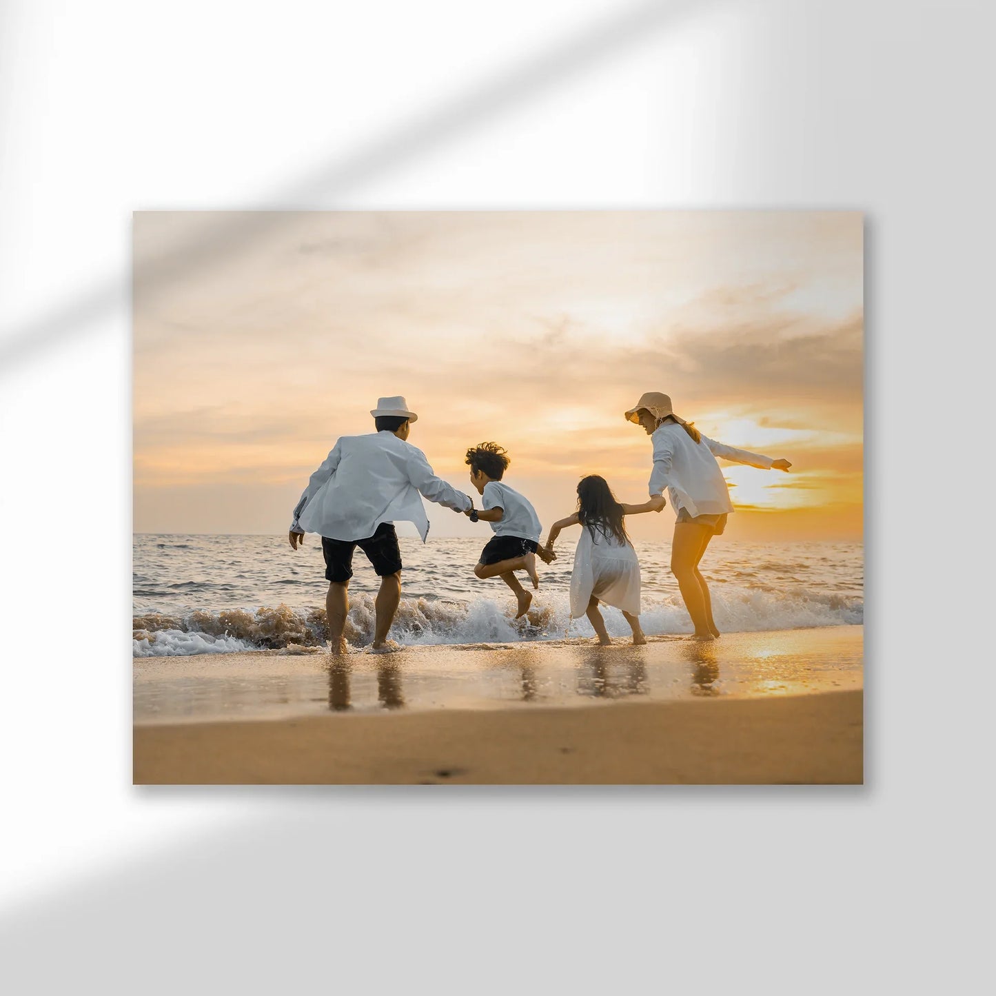 Mounted print featuring family playing at the beach during sunset, warm tones and horizontal layout, frameless display with soft shadow on wall.
