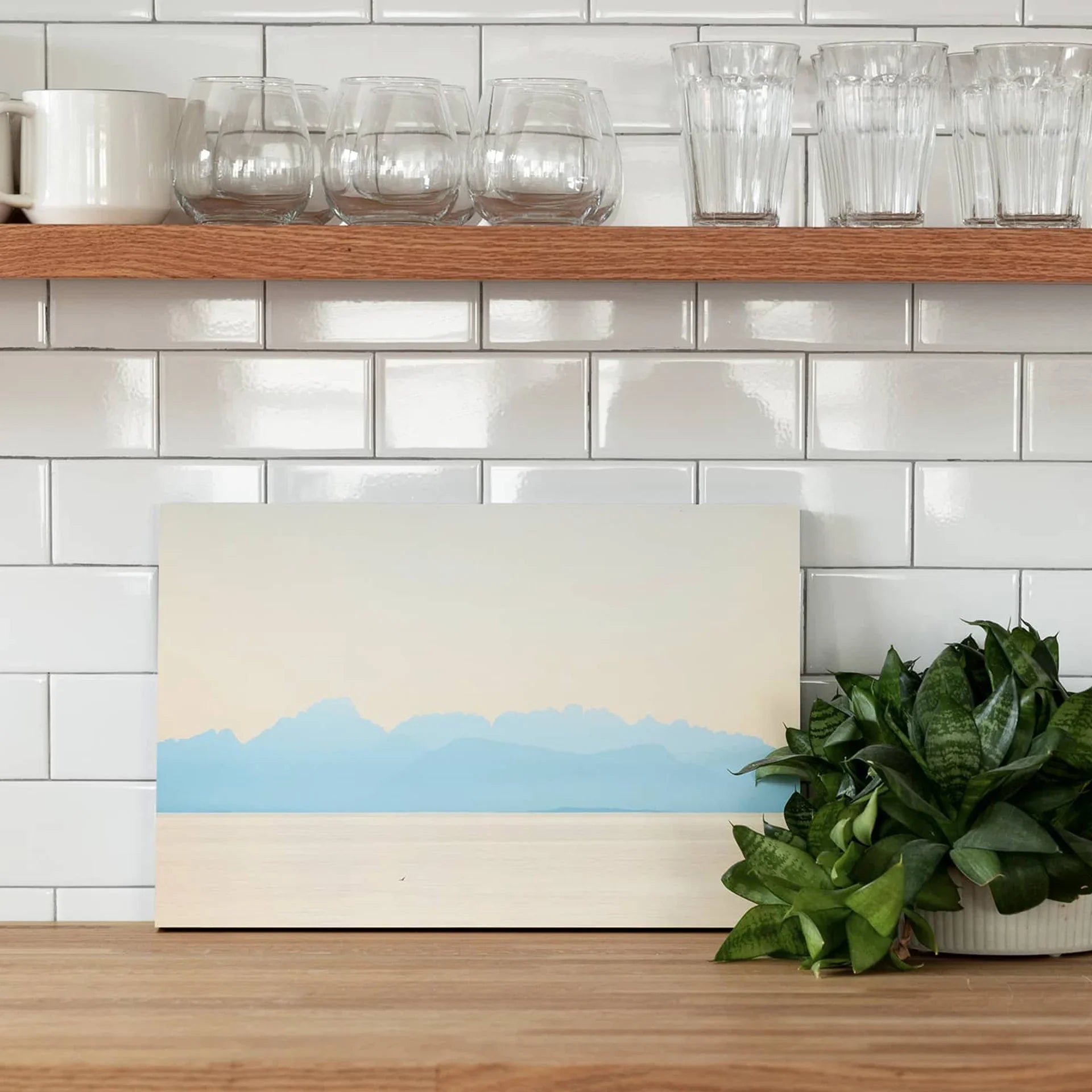 Mounted photo print featuring a minimalist mountain landscape in soft blue tones, displayed against white subway tile with glassware above.