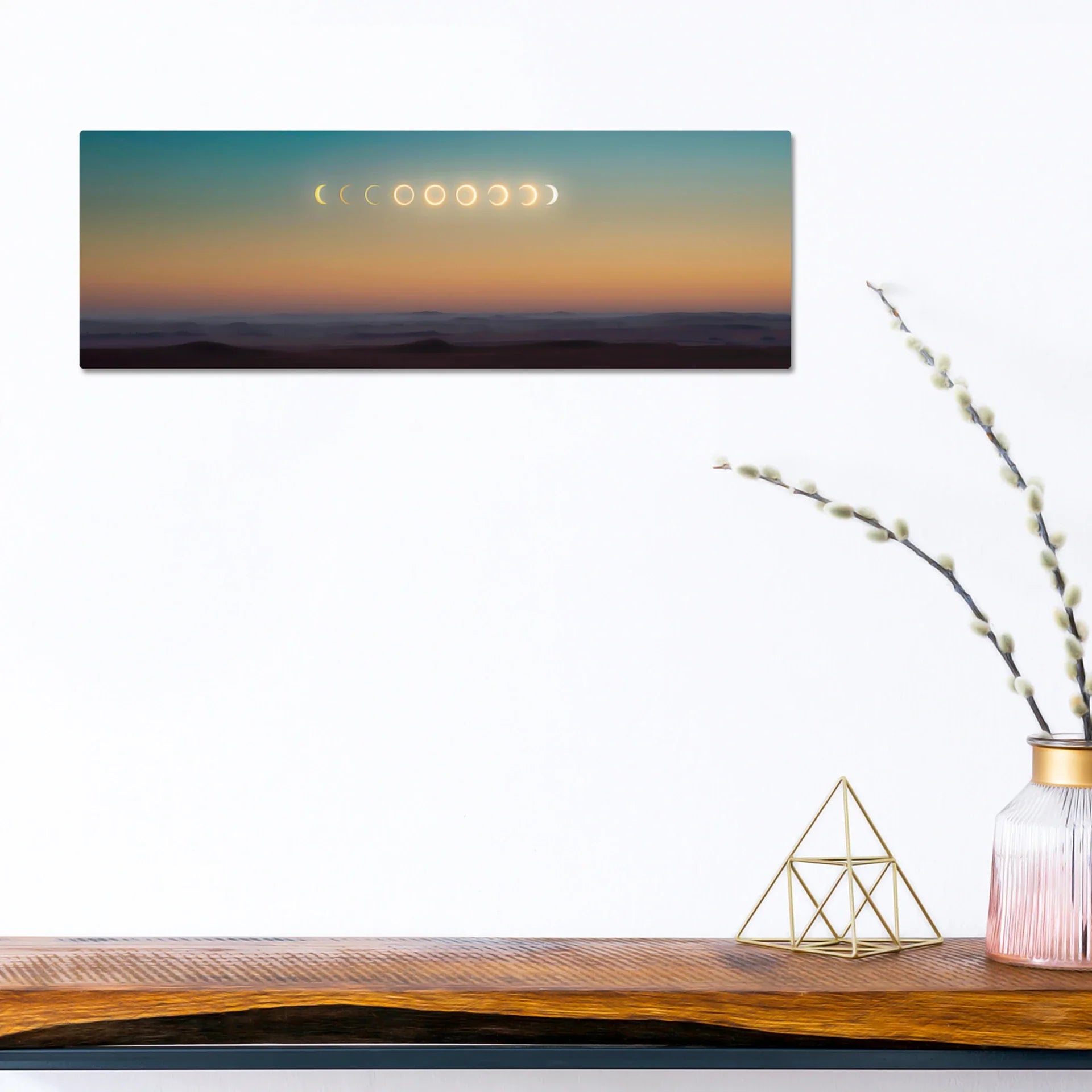 Panoramic print featuring moon phases across a gradient sunset sky above layered mountain silhouettes, mounted on a white wall above a wooden console table.