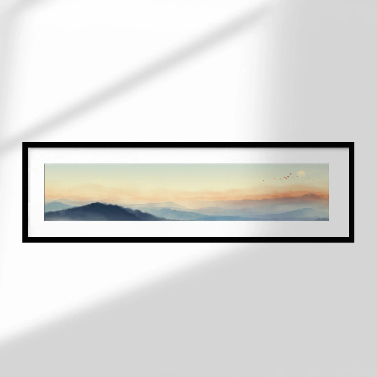 Framed panoramic print with white mat and black frame displaying a soft mountain landscape at sunrise, hung on a neutral wall.