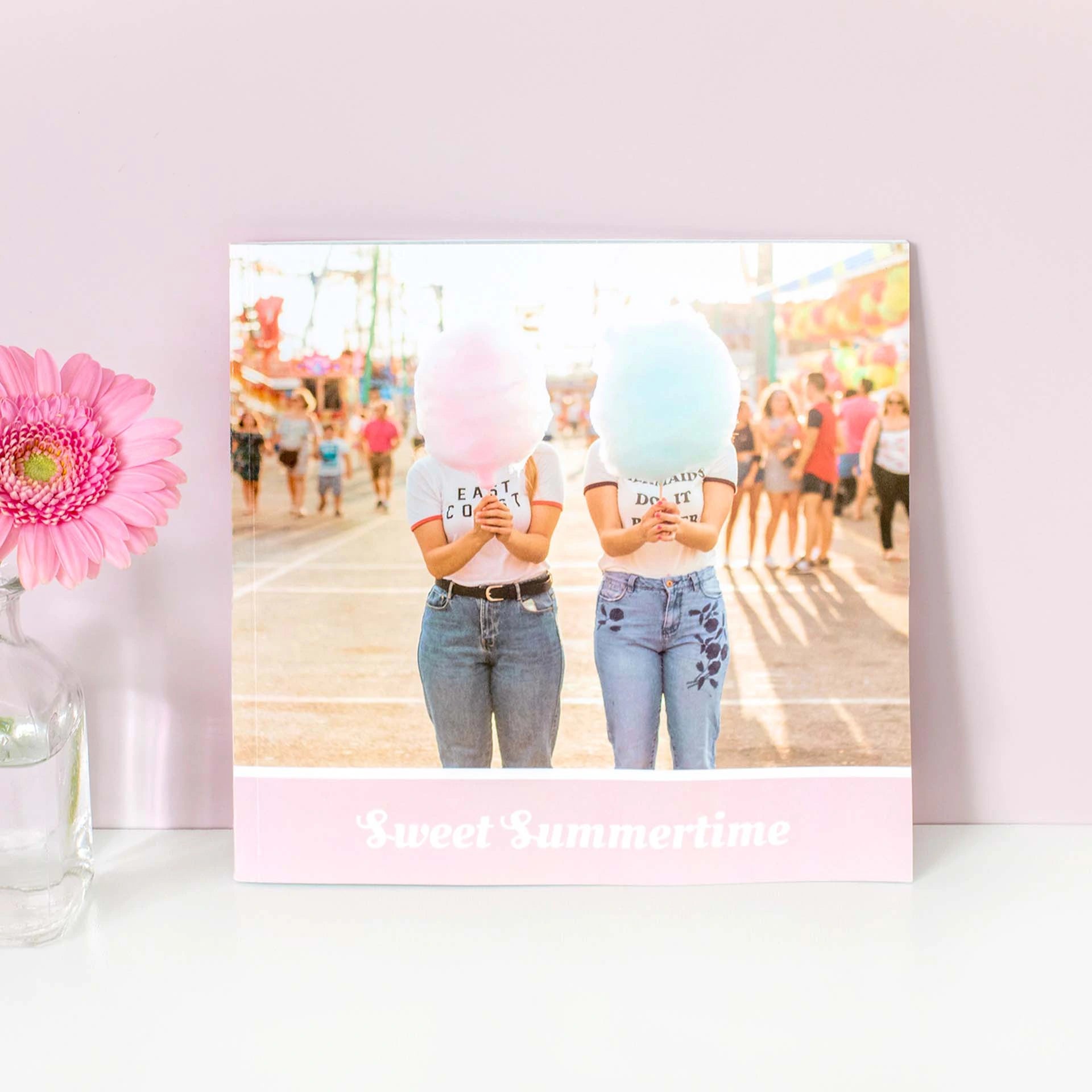 Softcover photo book standing upright with carnival scene on cover showing two people holding cotton candy, displayed on shelf with flower.