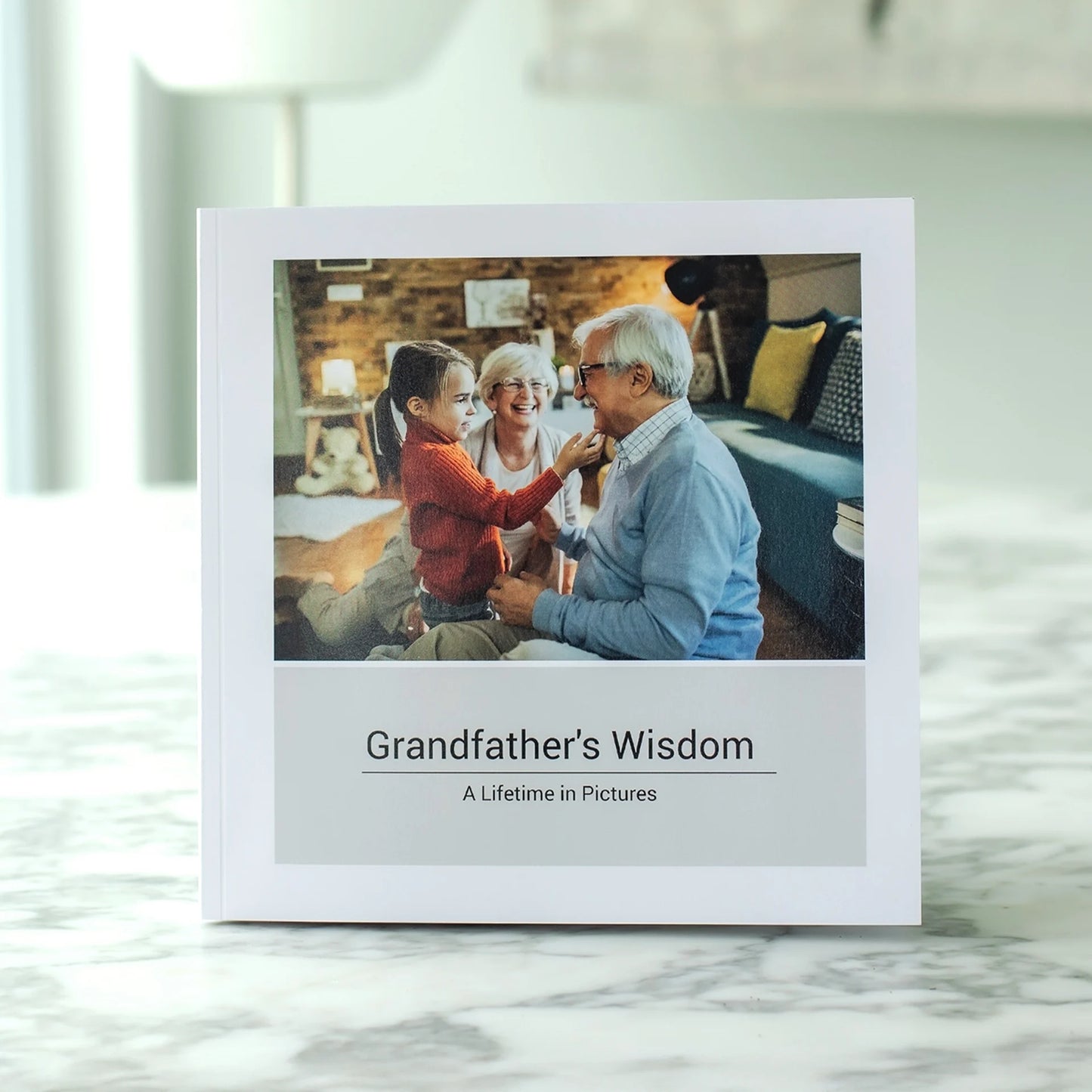 Softcover photo book titled 'Grandfather's Wisdom: A Lifetime in Pictures' standing upright on a marble surface, featuring a cover photo of a grandfather and grandmother laughing with their young granddaughter indoors
