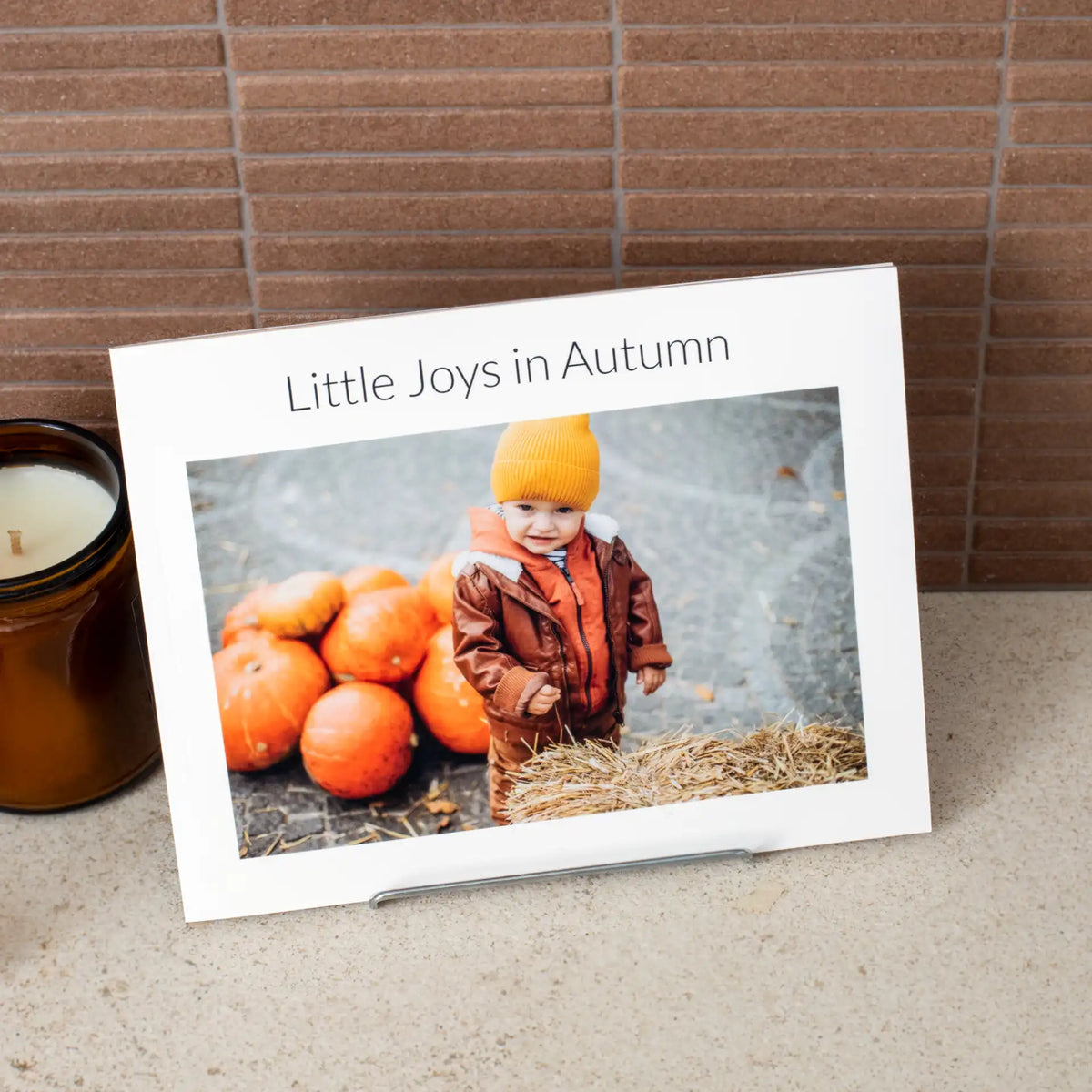 Single softcover photo book page titled 'Little Joys in Autumn,' showing a toddler in a beanie standing beside a pile of pumpkins.
