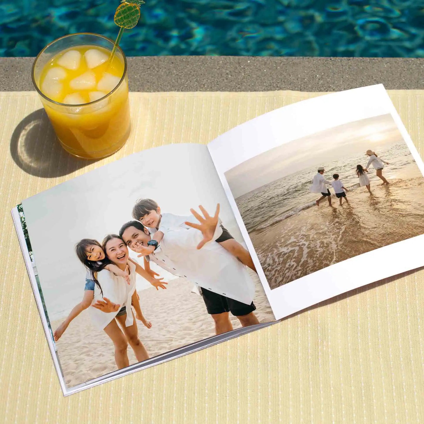Softcover photo book open to beach family photos including group portrait and children playing by shoreline, displayed beside drink near pool.