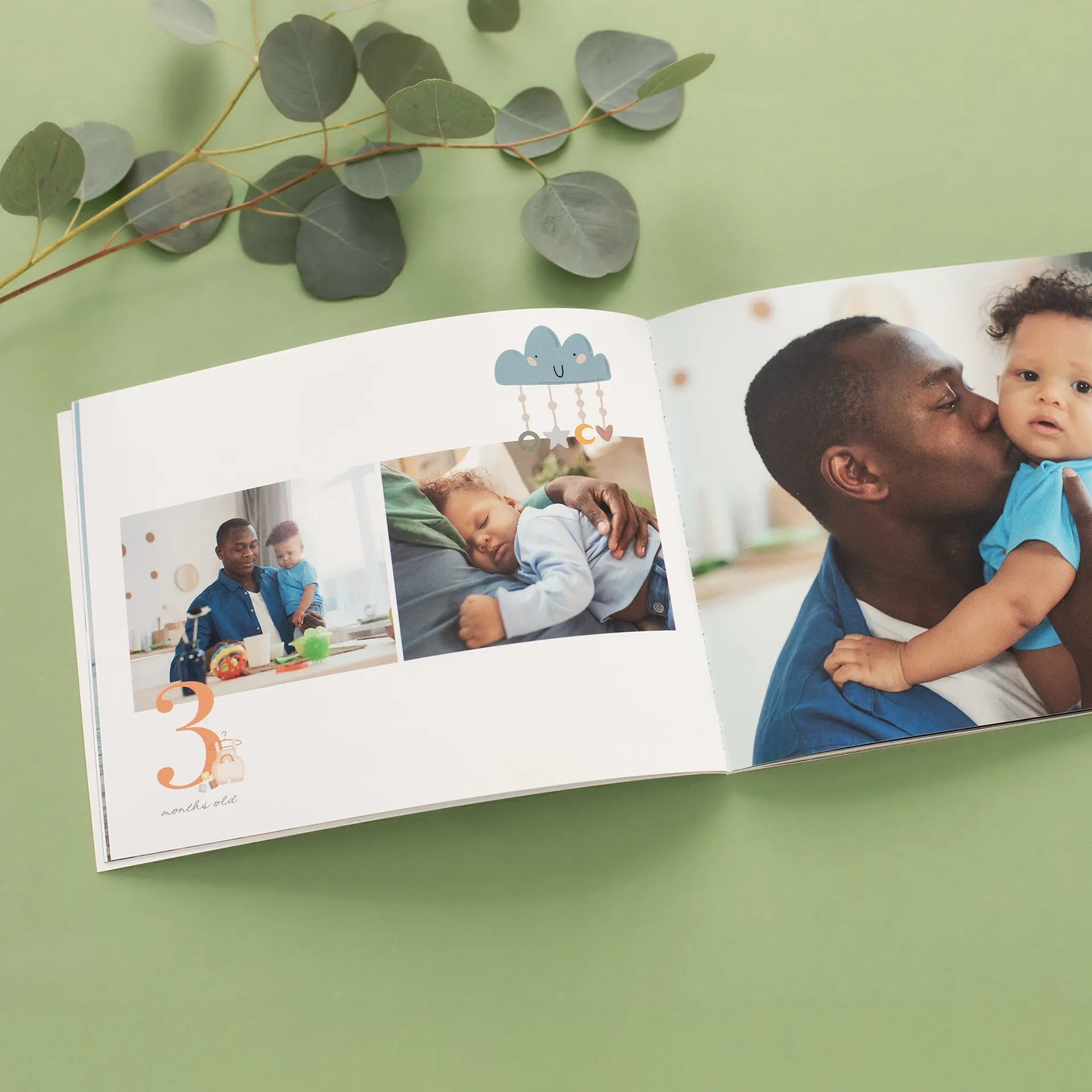 Open softcover photo book on a green surface displaying a baby milestone spread labeled '3 months old' with playful cloud stickers, showing a father bonding with his infant son in multiple photos