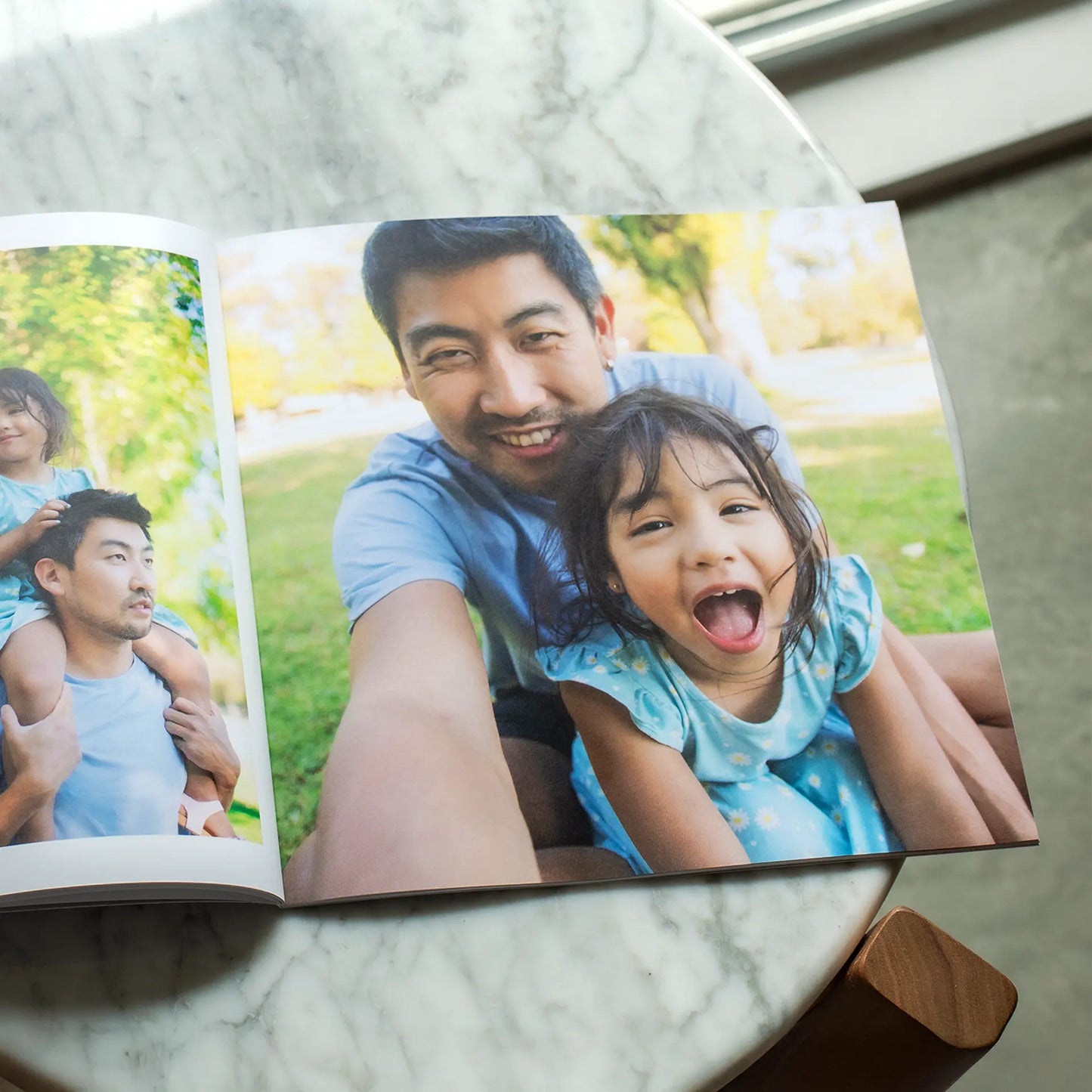 Open softcover photo book resting on a marble table showing a joyful spread of a father and young daughter laughing together outdoors in a sunny park