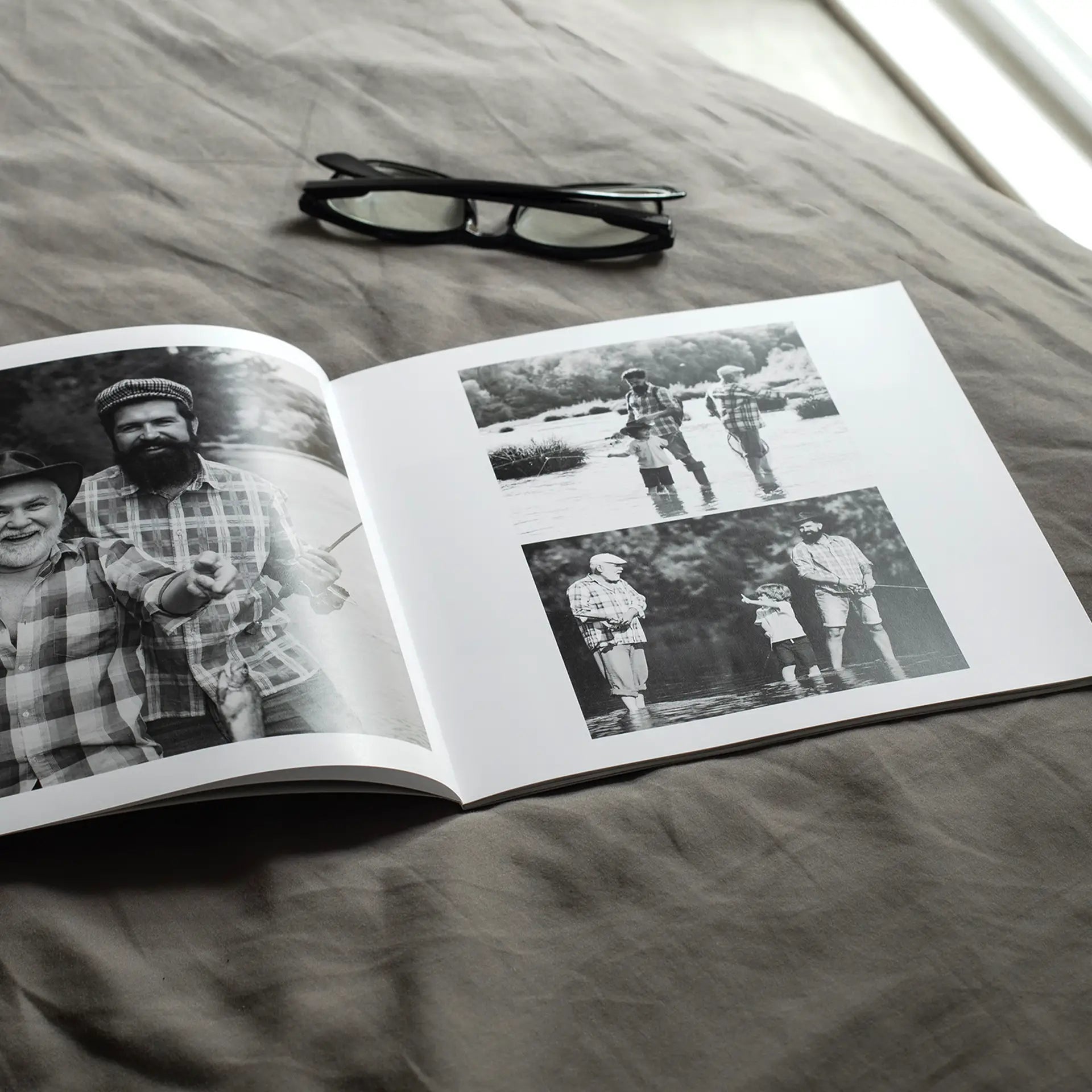 Open softcover photo book on a grey linen surface beside a pair of glasses, showing a black and white spread of a father, grandfather, and young boy fishing together in a river