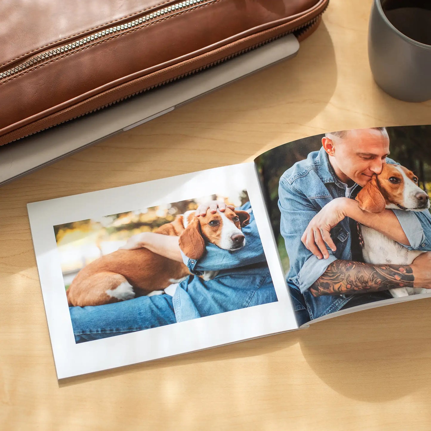 Open softcover photo book on a wooden desk beside a leather bag and coffee mug, displaying two photos of a man affectionately hugging his beagle outdoors