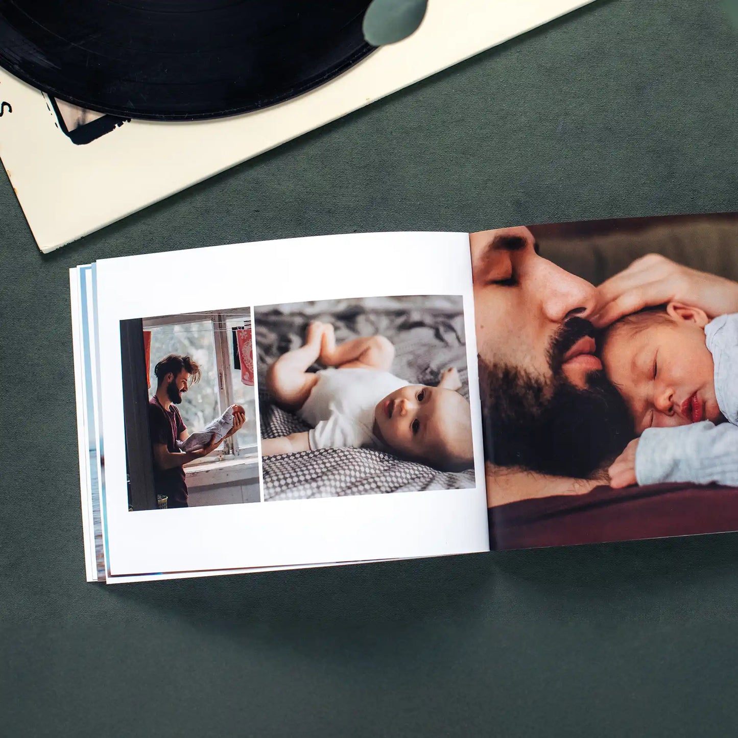 Open softcover photo book on a dark green surface beside a vinyl record, showing a tender collage spread of a bearded father caring for and cuddling his newborn baby
