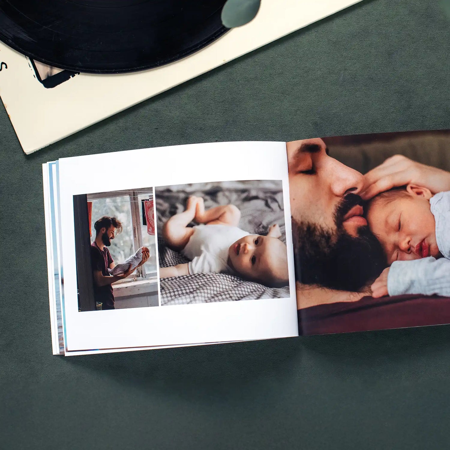 Open softcover photo book on a dark green surface beside a vinyl record, showing a tender collage spread of a bearded father caring for and cuddling his newborn baby