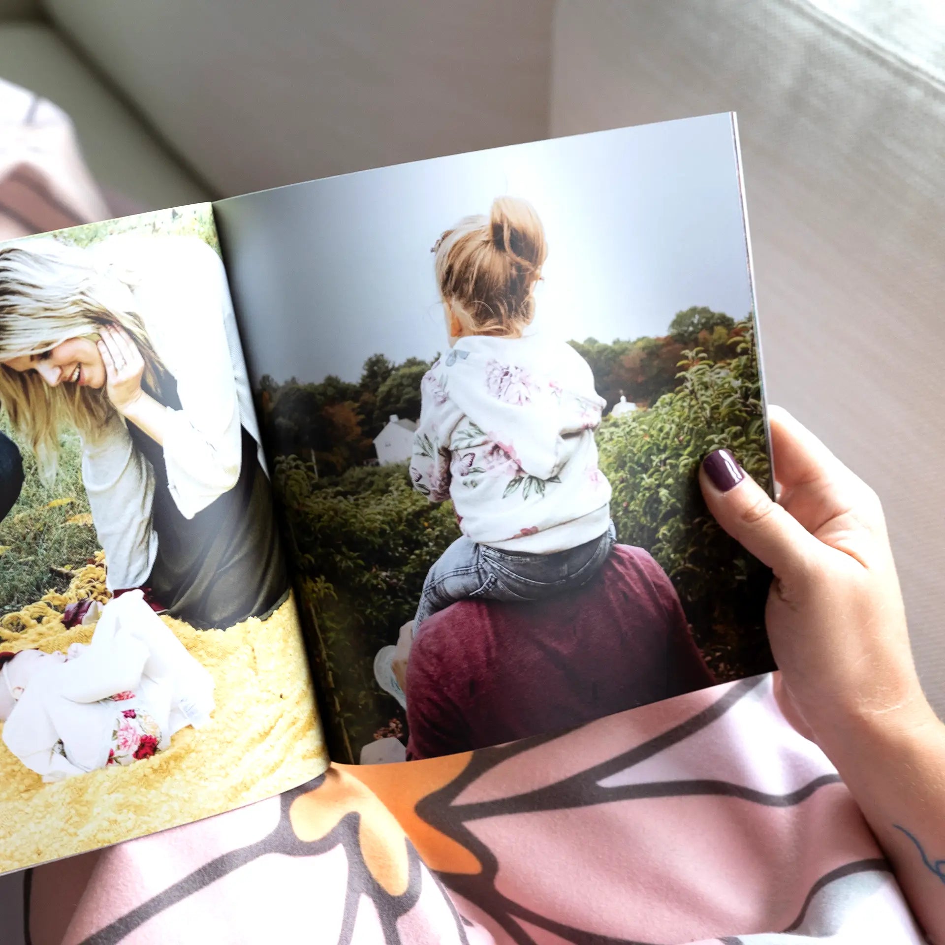 A person holding open a softcover photo book displaying a family spread with a photo of a mother laughing outdoors and a full-page photo of a young child sitting on a parent's shoulders in a garden
