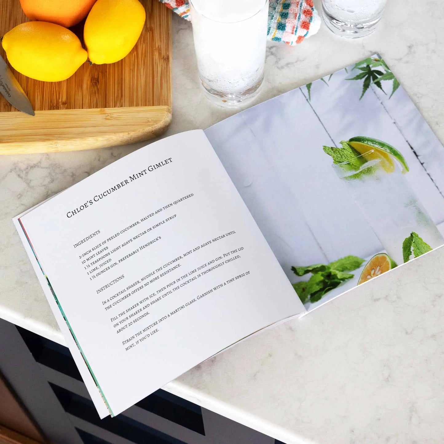 Softcover photo book open to recipe-style layout with text on one page and cocktail image on opposite page, placed on kitchen countertop.