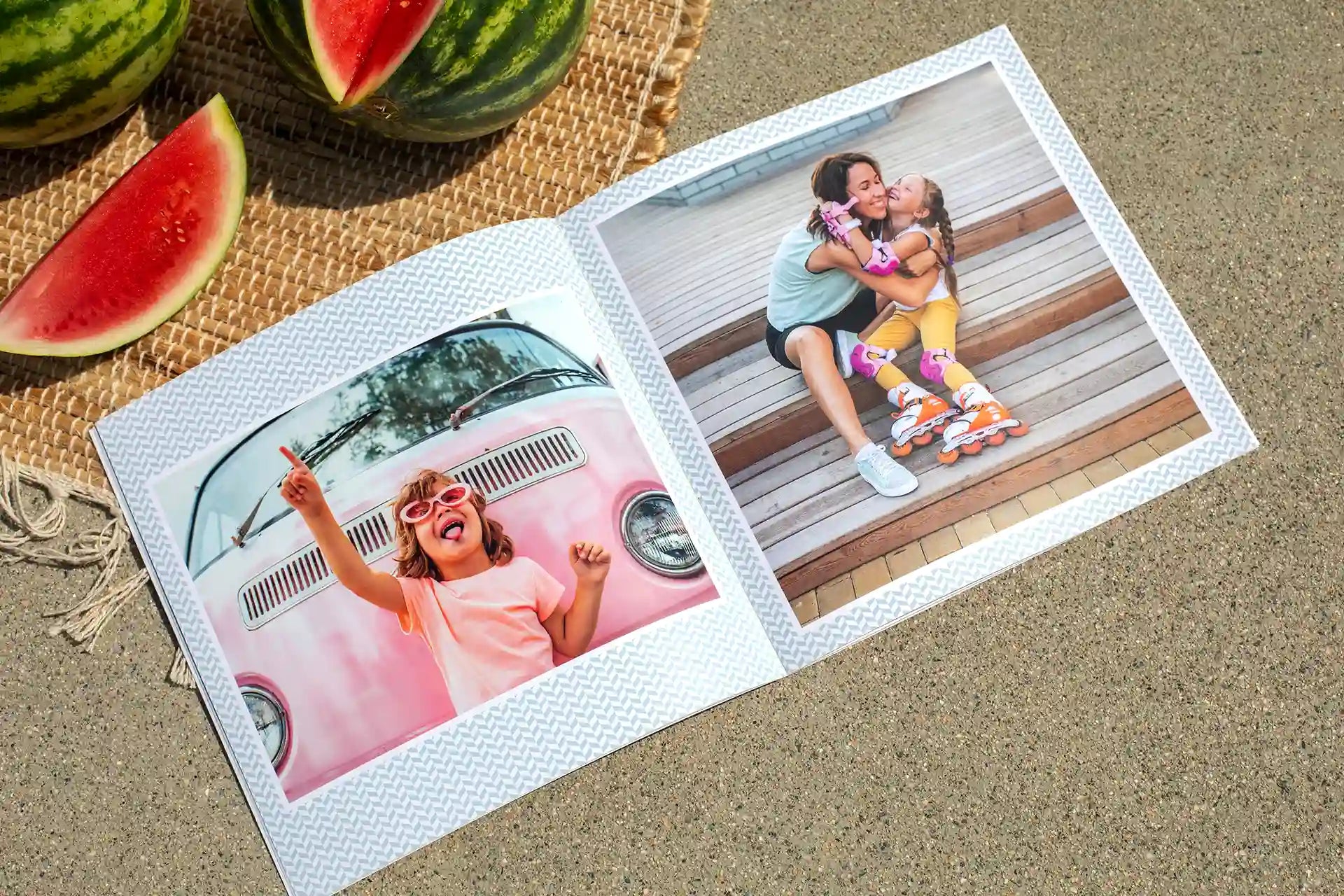 Open softcover photo book with a chevron-patterned border layout, showing two summer photos — a young girl posing with sunglasses in front of a pink van, and a mother hugging her daughter on outdoor steps — displayed on a sandy surface beside sliced watermelon.