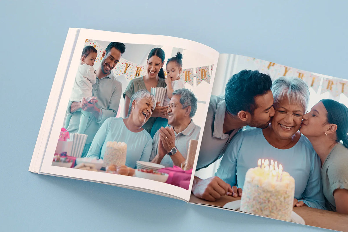Open softcover photo book showing a birthday celebration: on the left, family members laughing together with birthday banners and party snacks in the background; on the right, grandparents being kissed on the cheek by their adult children in front of a lit birthday cake.