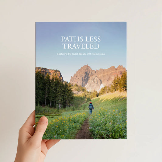 8.5x11 softcover photo book held in one hand, featuring a mountain landscape cover with a person hiking along a narrow trail.