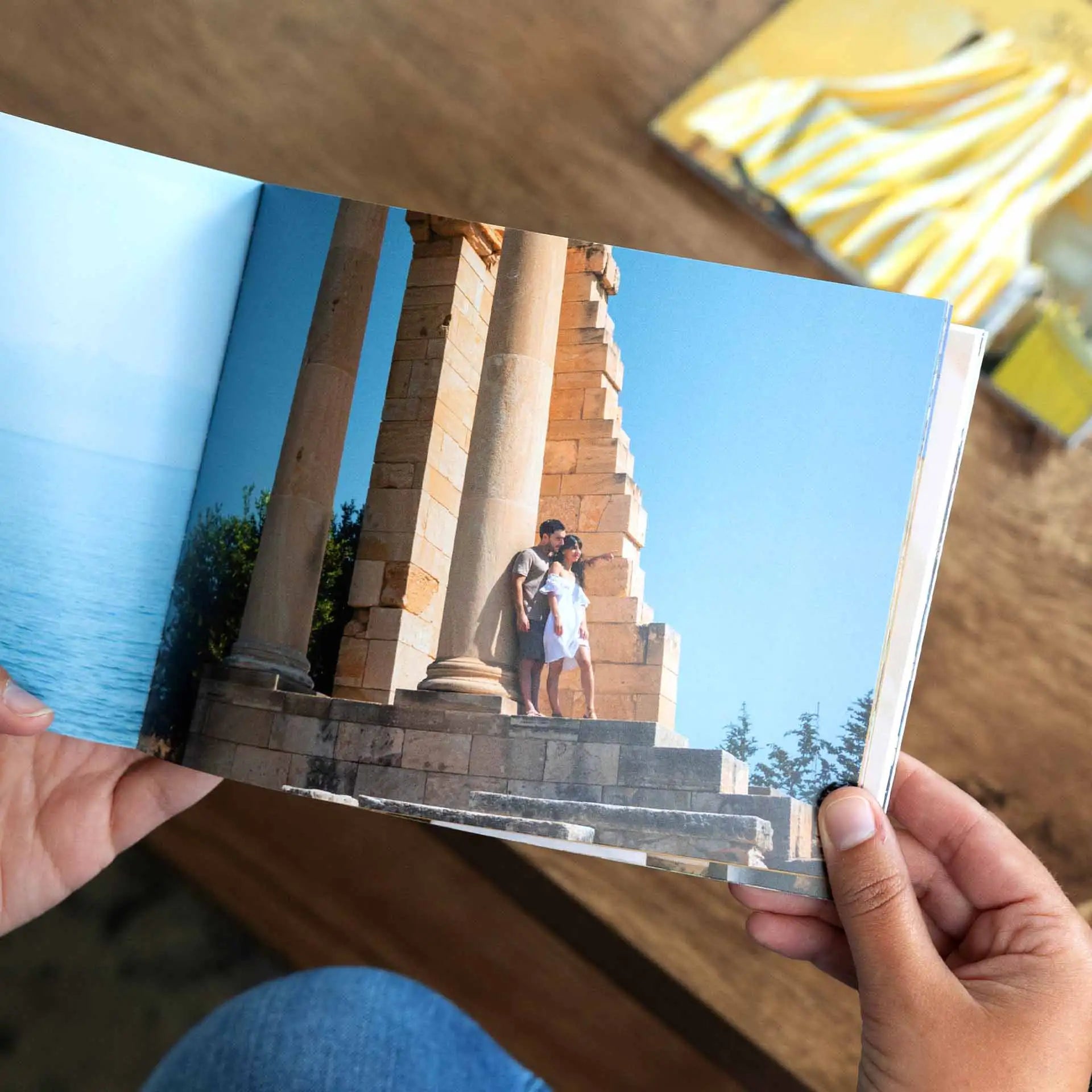 Softcover photo book held open showing full-page image of couple standing near stone columns outdoors, printed across two-page spread.