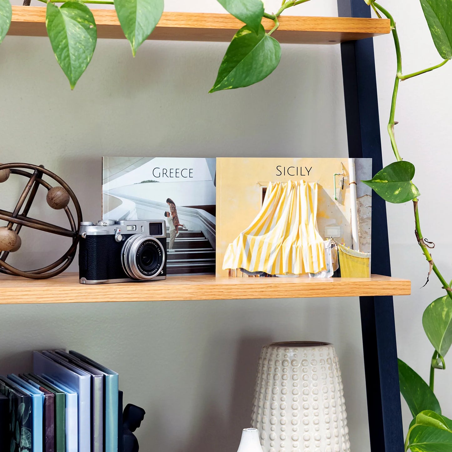 Two softcover photo books displayed on a wooden shelf, showing travel-themed covers labeled Greece and Sicily beside a camera and home decor