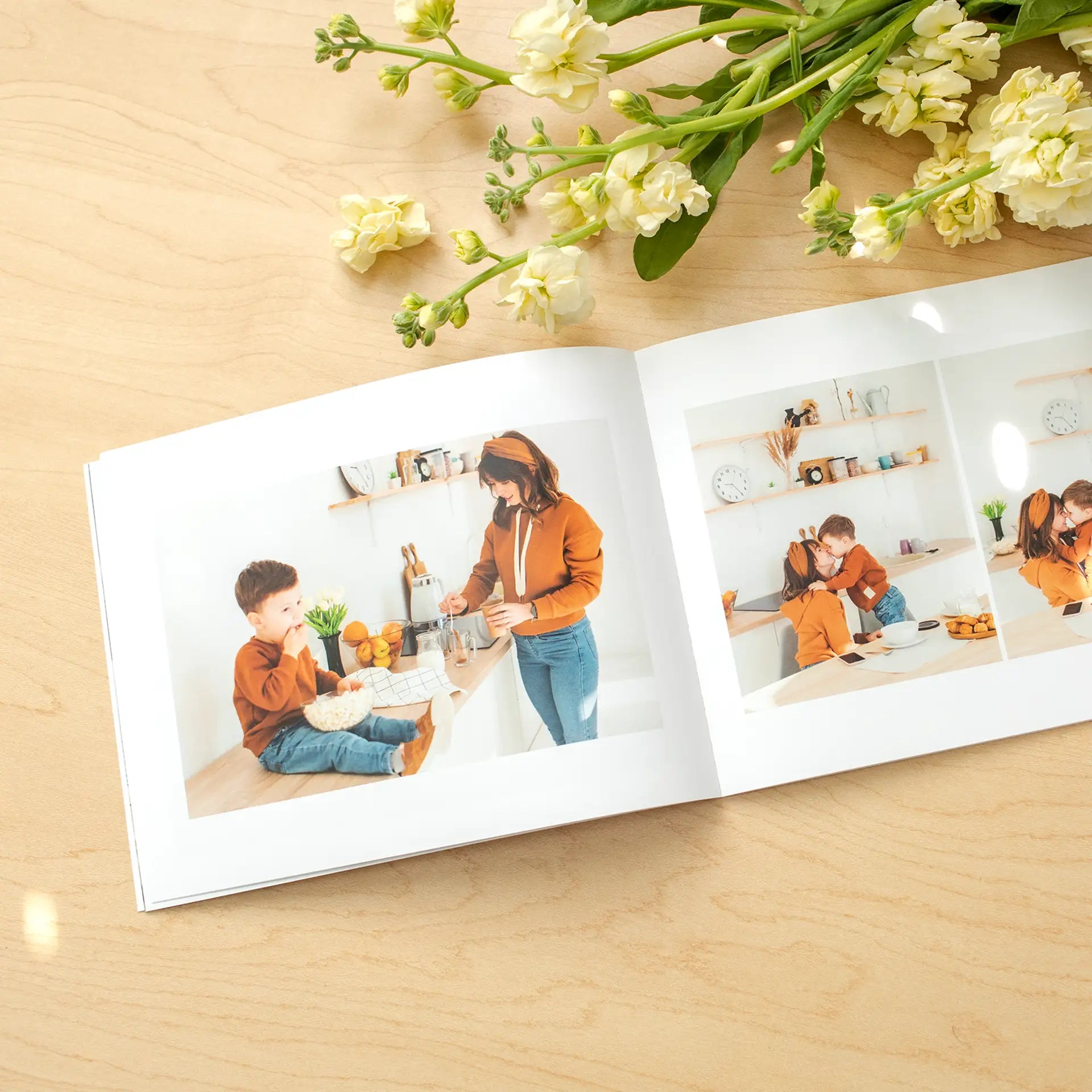 Softcover photo book open to multi-image collage of a person and child in a bright kitchen setting, arranged in clean grid layout with flowers above.