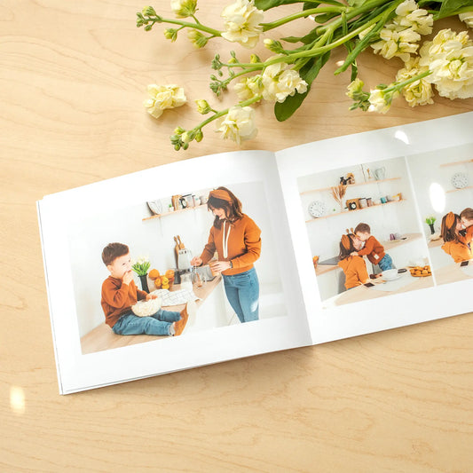 Softcover photo book open to multi-image collage of a person and child in a bright kitchen setting, arranged in clean grid layout with flowers above.