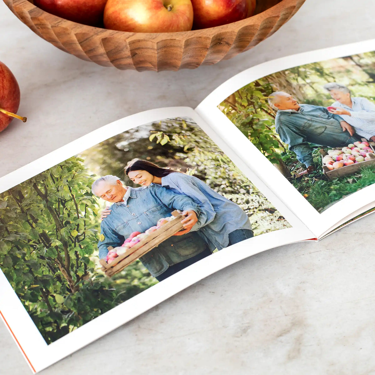 Softcover photo book open to fall photos of a family picking apples together in an orchard, shown beside a bowl of apples.