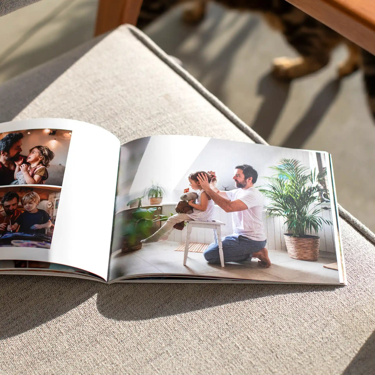 Softcover photo book open to a full-page photo of a father fixing his daughter's hair indoors, with smaller photos of family moments.