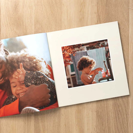 Softcover photo book open to a warm hug between mother and daughter, facing a photo of a child painting outside in autumn light.
