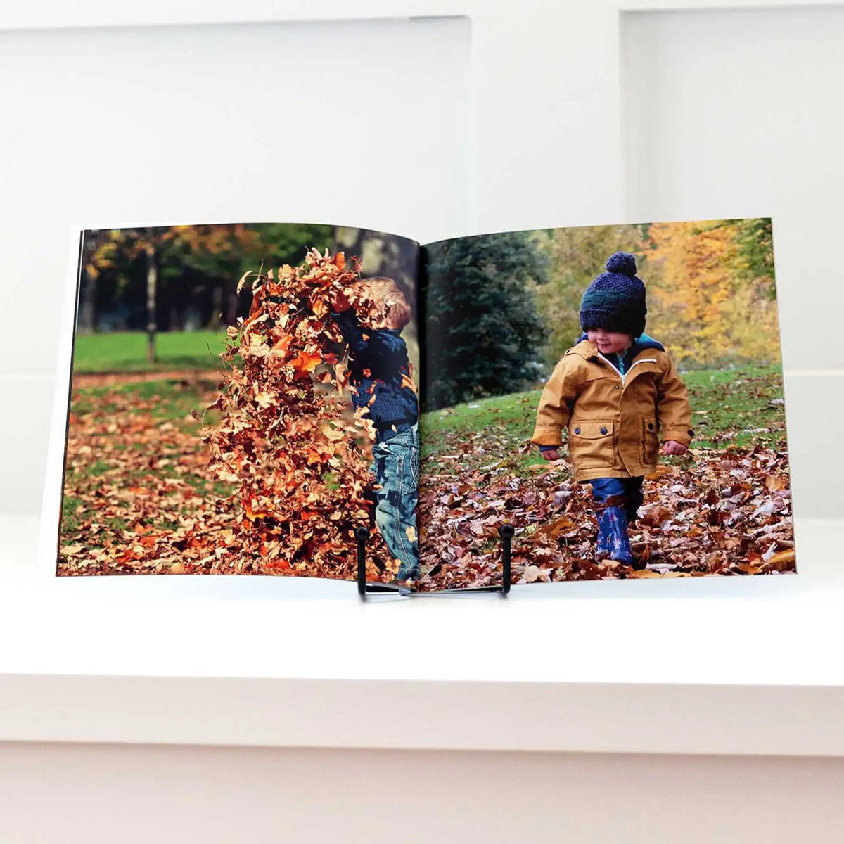 Softcover photo book standing upright, open to photos of children playing and walking through piles of autumn leaves.