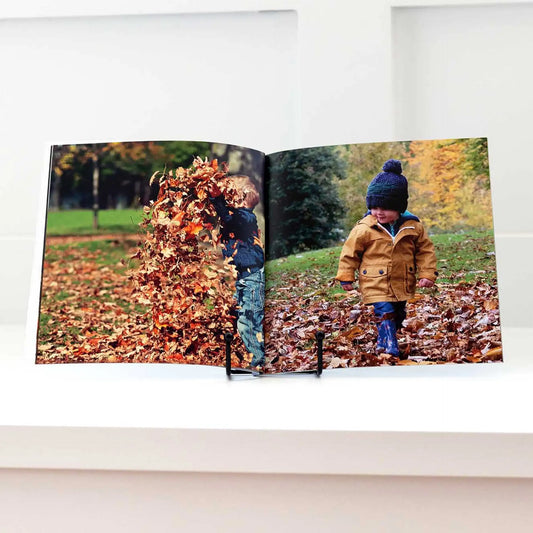 Softcover photo book standing upright, open to photos of children playing and walking through piles of autumn leaves.