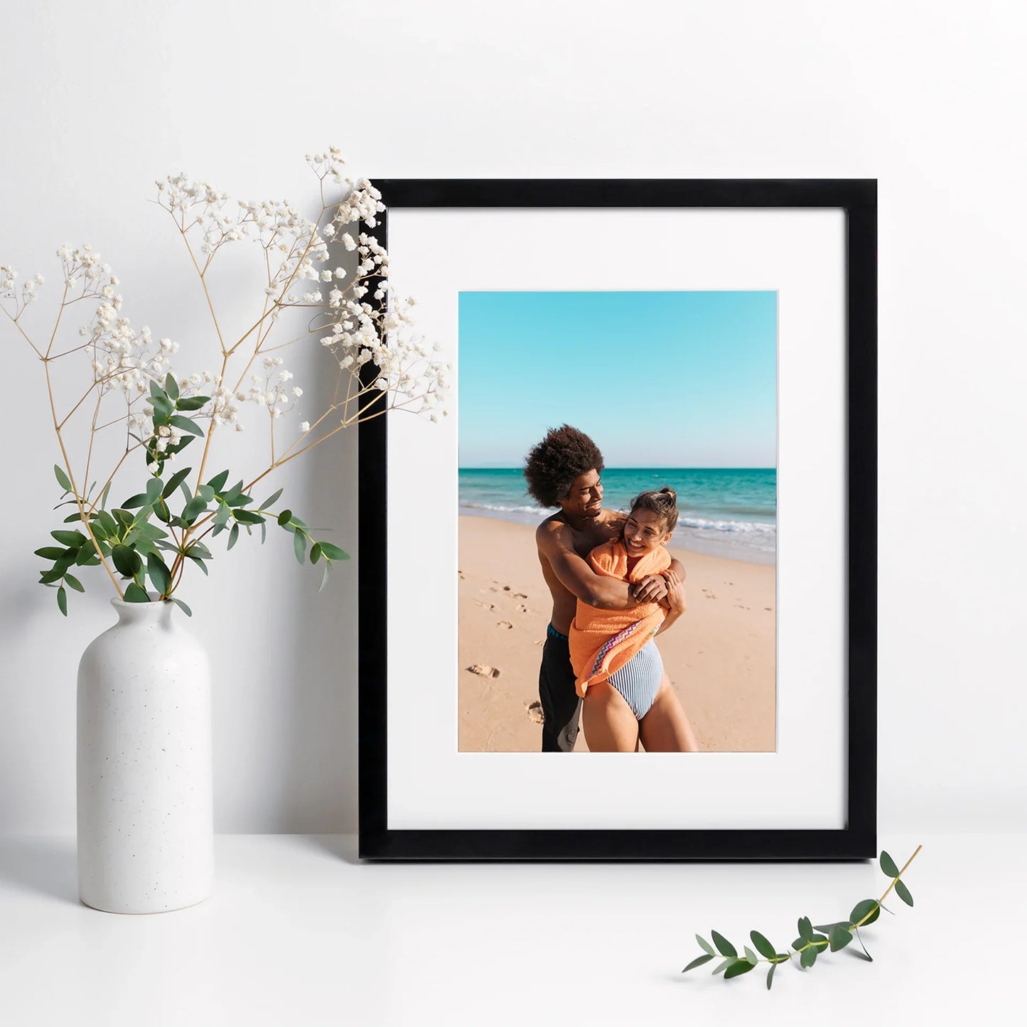 Black framed vertical photo print of a couple embracing on a sandy beach with turquoise ocean background, displayed beside a white vase with greenery.