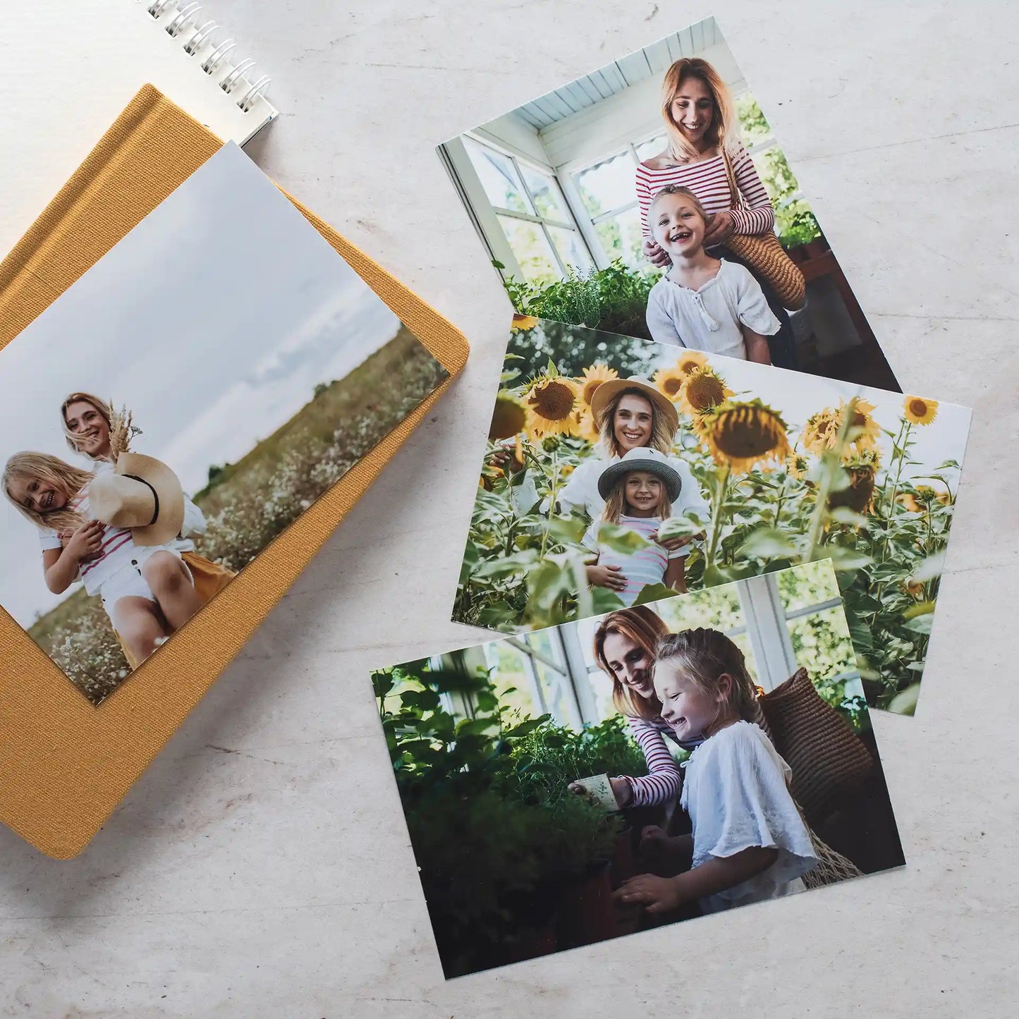 Stack of glossy family photo prints beside a mustard fabric journal, showing a mother and child indoors and in a sunflower field.