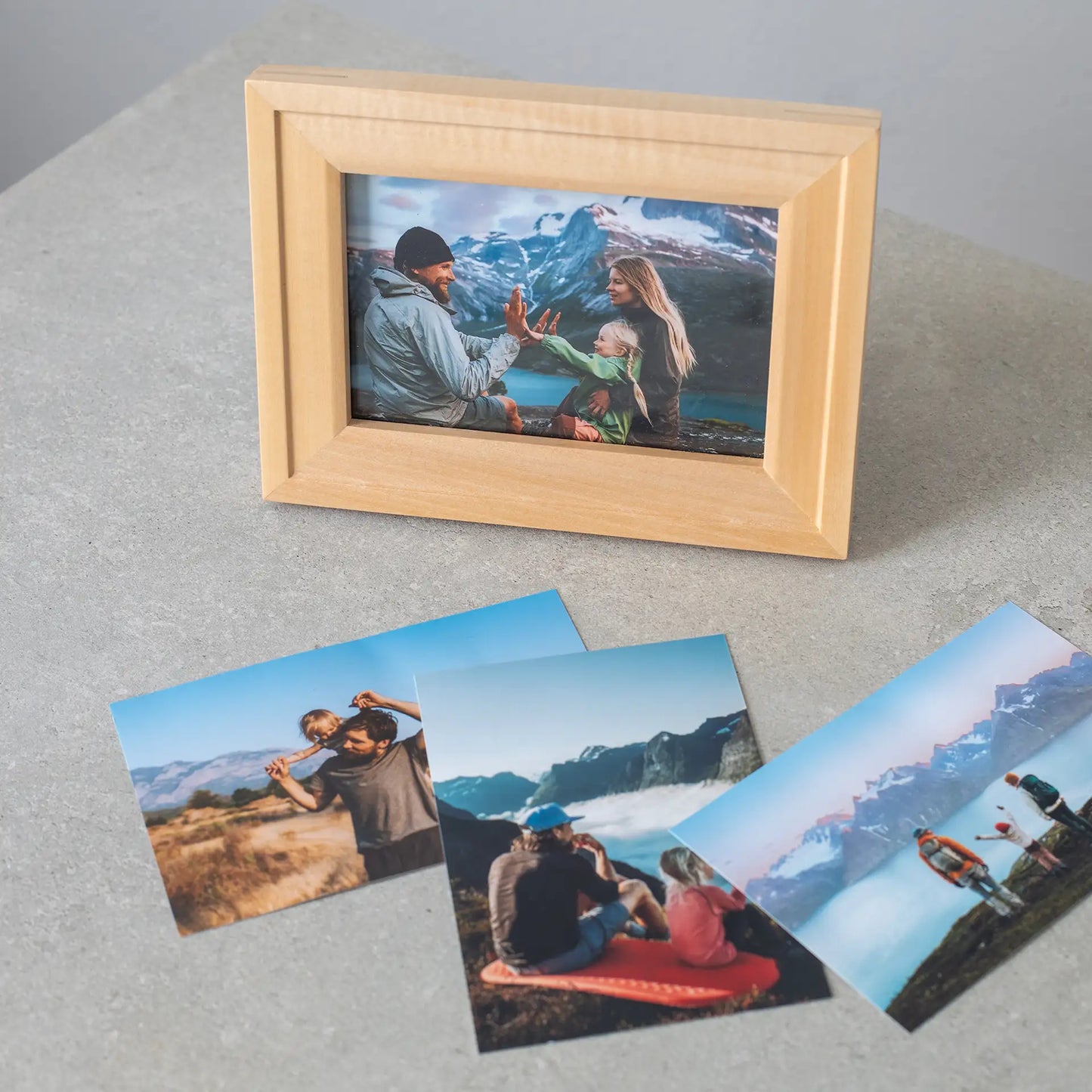 Horizontal wooden framed family photo of two adults and a child high-fiving with mountains and a lake in the background, displayed on a tabletop.