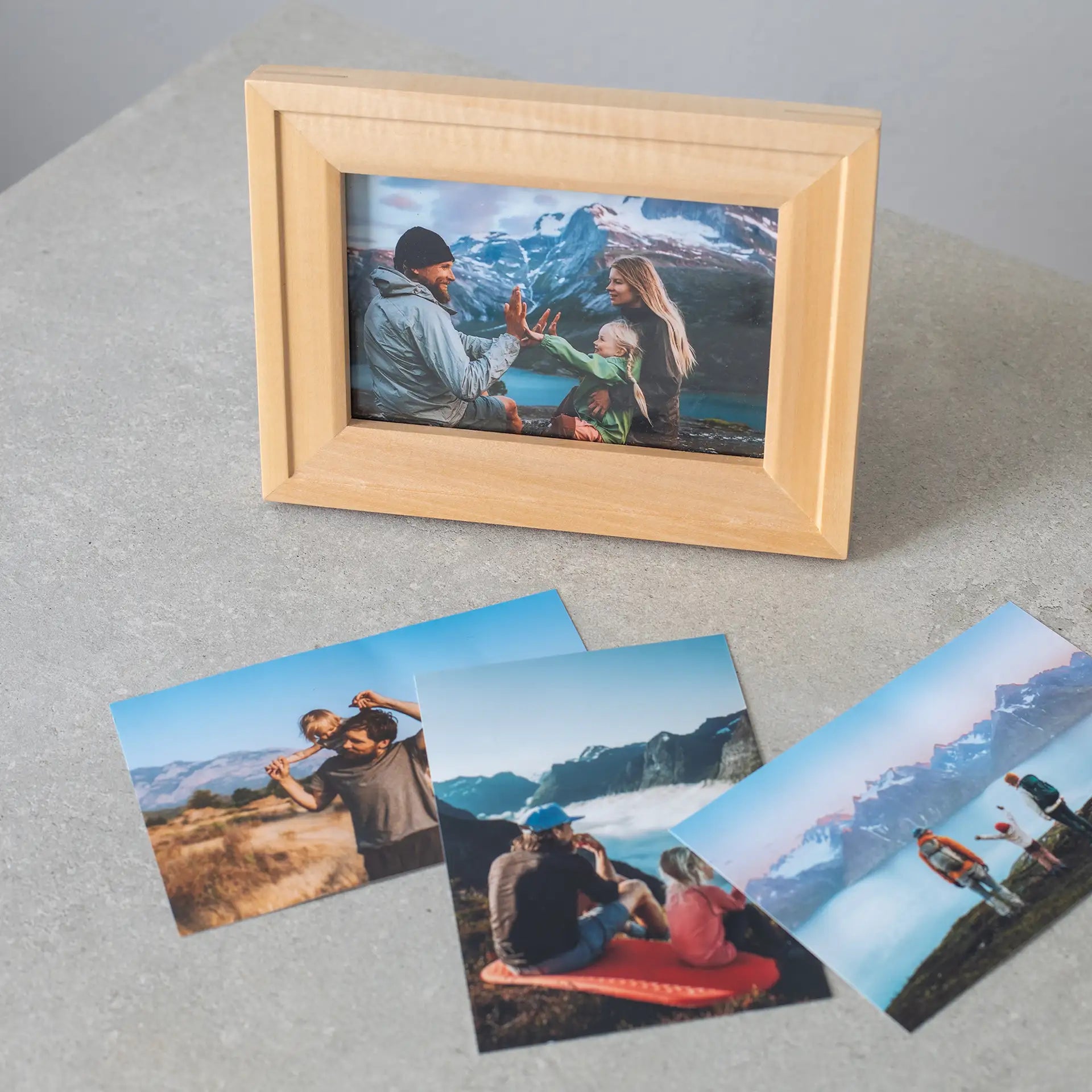 Horizontal wooden framed family photo of two adults and a child high-fiving with mountains and a lake in the background, displayed on a tabletop.